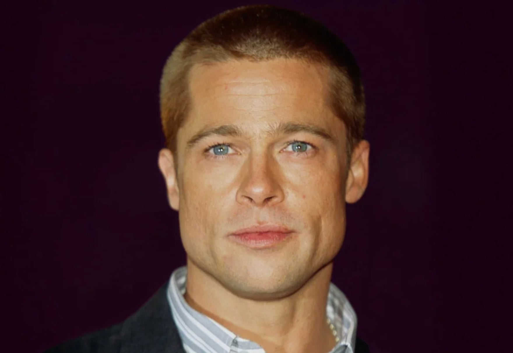 A man with short blond hair and blue eyes is wearing a collared shirt and dark jacket, looking slightly past the camera against a dark purple background.