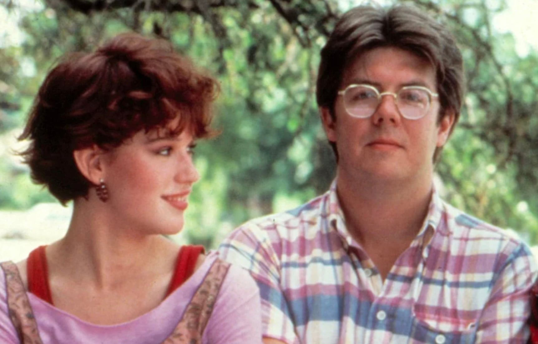 A young woman with short red hair wearing a pink top smiles and looks at a man in glasses and a plaid shirt who faces the camera, with green trees in the background.