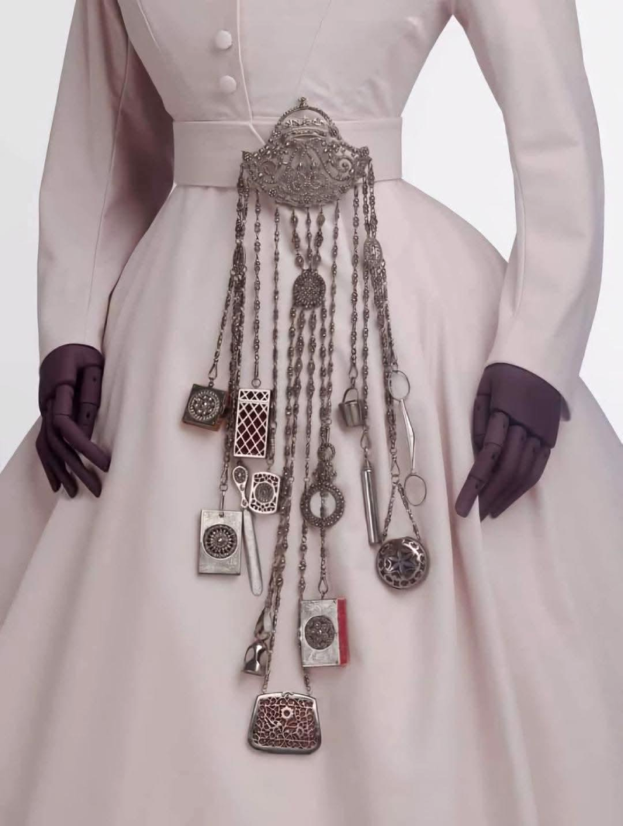 A mannequin in a pale, long-sleeved dress wears a decorative chatelaine belt with multiple ornate silver chains holding vintage tools and accessories, such as scissors, keys, and small boxes.