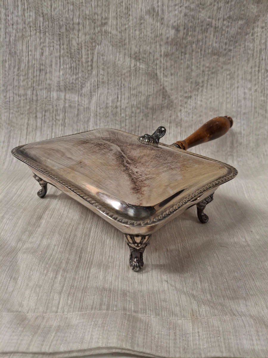 A vintage silver-plated serving dish with a wooden handle, ornate legs, and a hinged lid, displayed on a light textured fabric background.