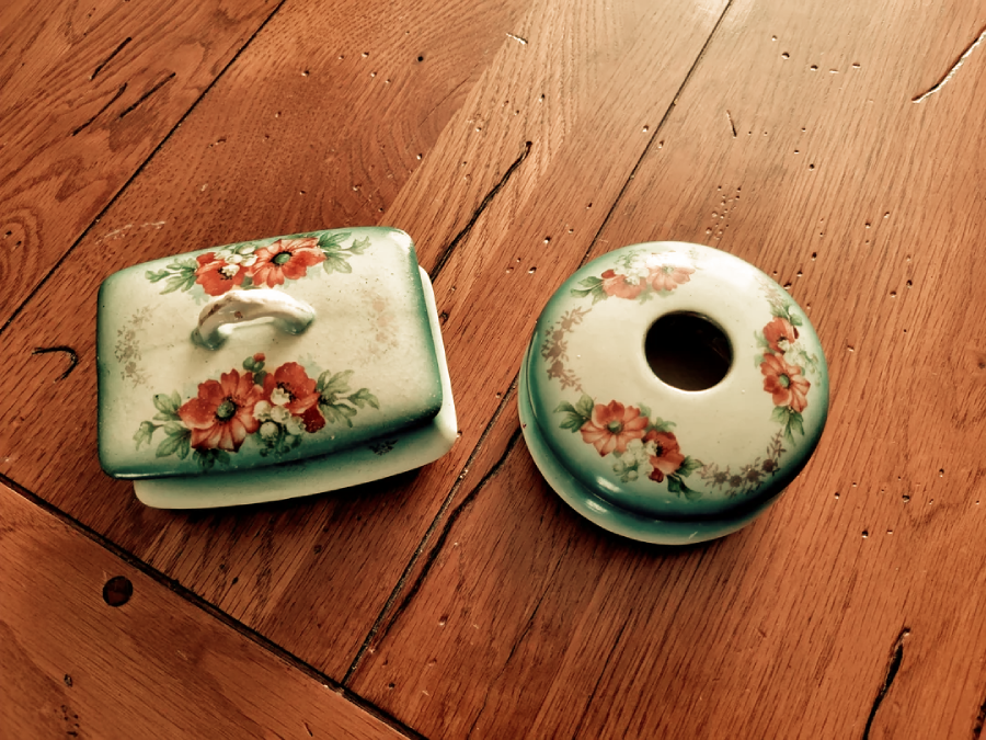 Two vintage porcelain items with floral patterns sit on a wooden table; one is a rectangular covered dish, and the other is a round holder with a large central hole, both decorated with red flowers and green leaves.