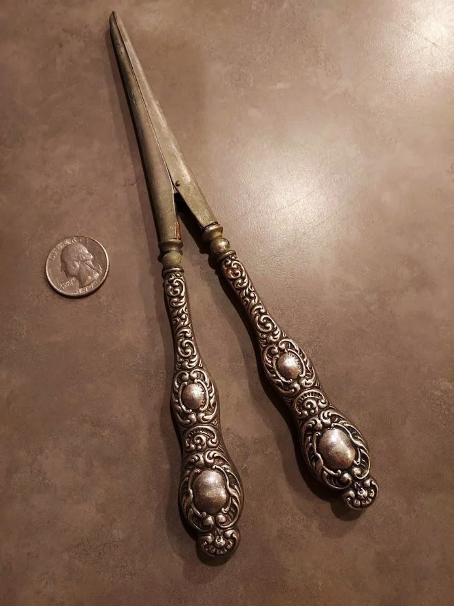 A pair of ornate, vintage metal tongs with detailed handles and pointed tips lying on a brown surface next to a U.S. quarter for size reference.