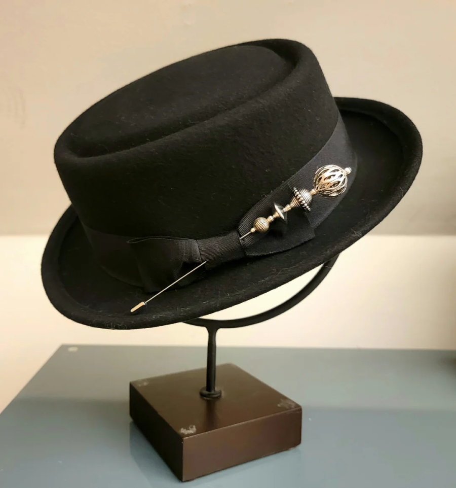 A black bowler hat with a decorative pin and ribbon band is displayed on a dark metal stand with a square base. The hat features a round crown and slightly curved brim.