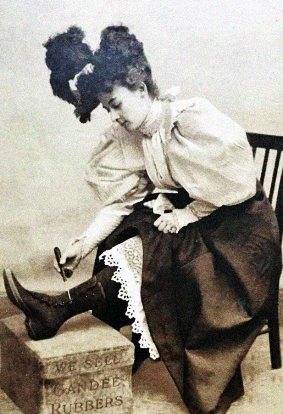 A woman in Victorian-era clothing uses a button hook to fasten buttons on her high-top boots, with one foot on a box labeled "WE SELL CANDY RUBBERS." She is seated on a wooden chair.