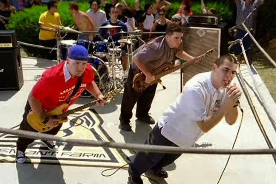 A band performs energetically outdoors on a small stage with guitars, drums, and microphones, surrounded by a crowd of enthusiastic fans in the background.