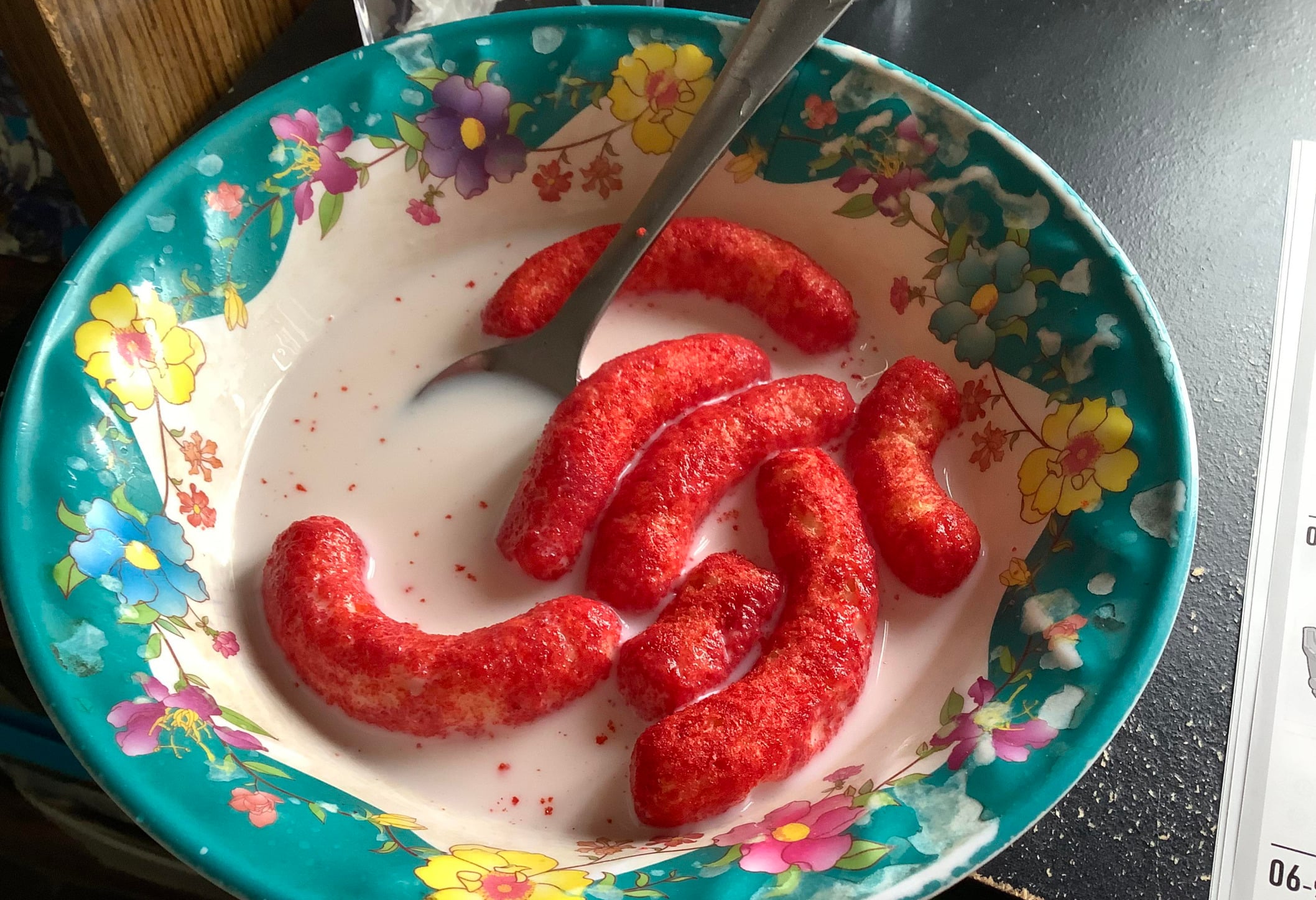 A colorful floral bowl filled with milk and several bright red, puffed snacks that resemble cheese puffs, with a spoon resting inside.