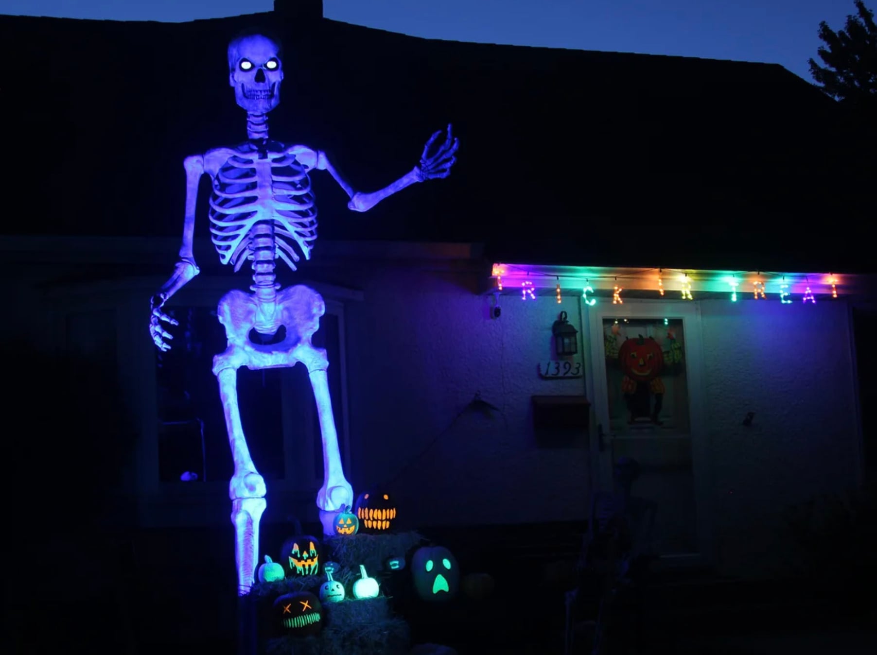 A large glowing skeleton Halloween decoration stands in a yard at night, surrounded by lit jack-o’-lanterns and multicolored string lights on a house in the background.