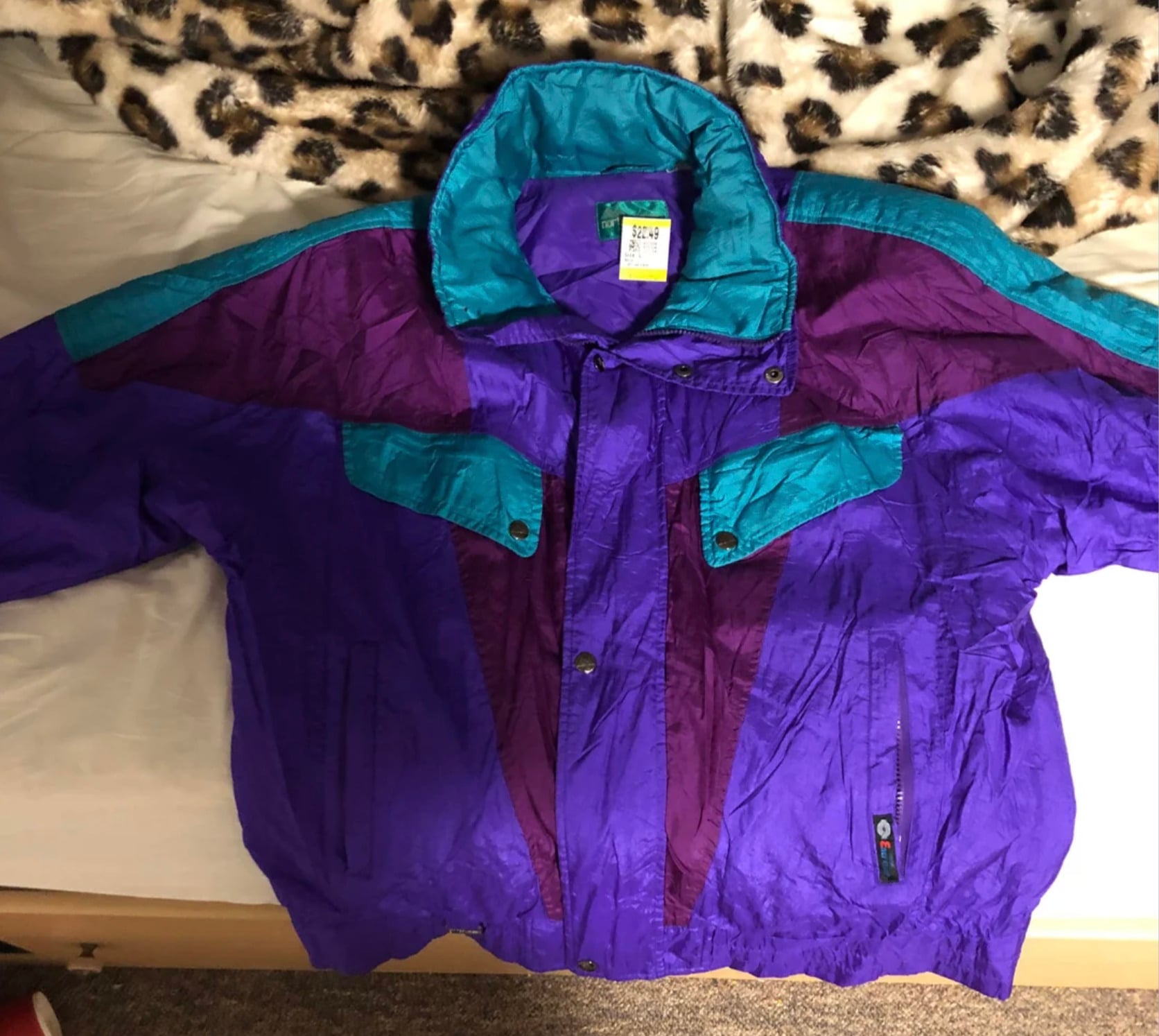 A vintage, color-block windbreaker jacket with purple, teal, and dark purple sections, laid flat on a bed with a leopard-print blanket. The jacket has front pockets and a high collar.