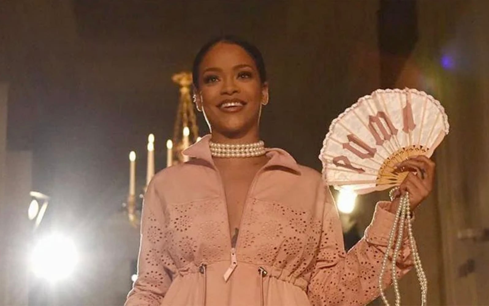 A woman wearing a light pink outfit and pearl necklace smiles while holding a pink hand fan with "PUMA" written on it. Bright lights and a chandelier are visible in the background.