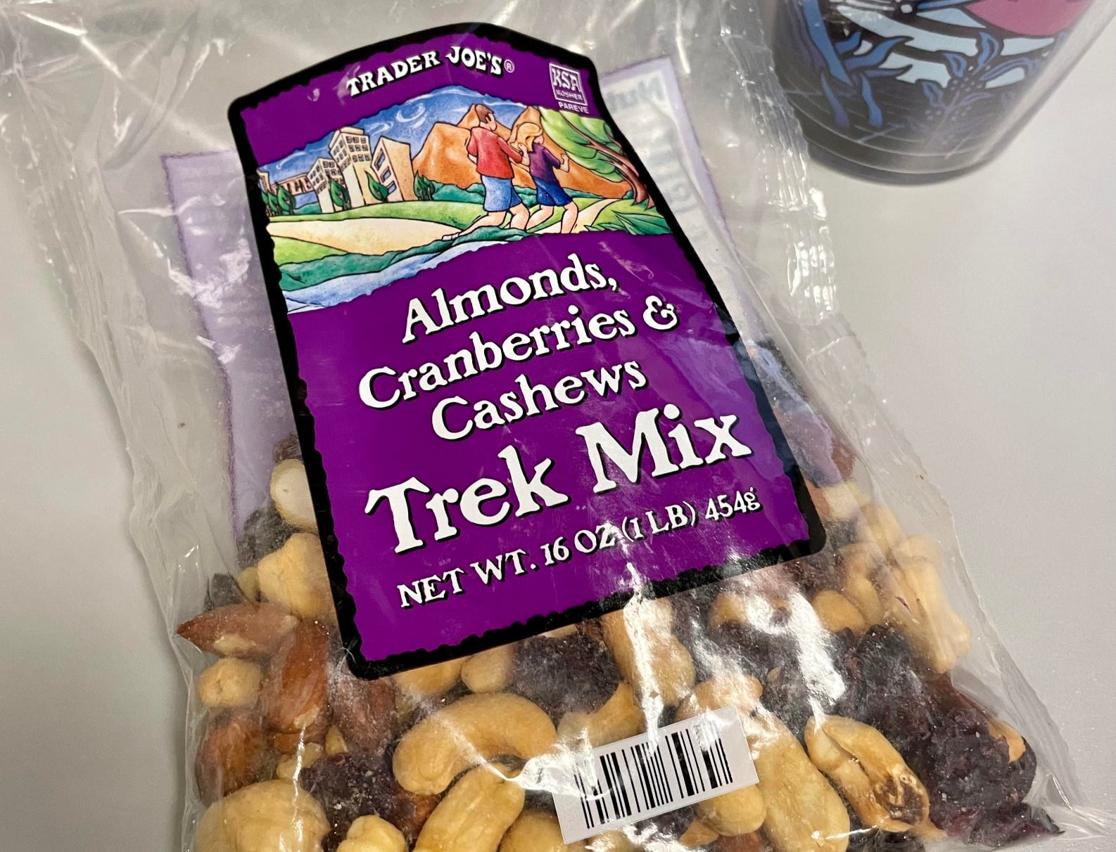 A partially opened bag of Trader Joe’s Trek Mix with almonds, cranberries, and cashews sits on a table. The purple label lists the net weight as 16 oz (1 lb) or 454 g. Some of the mix is visible through the packaging.