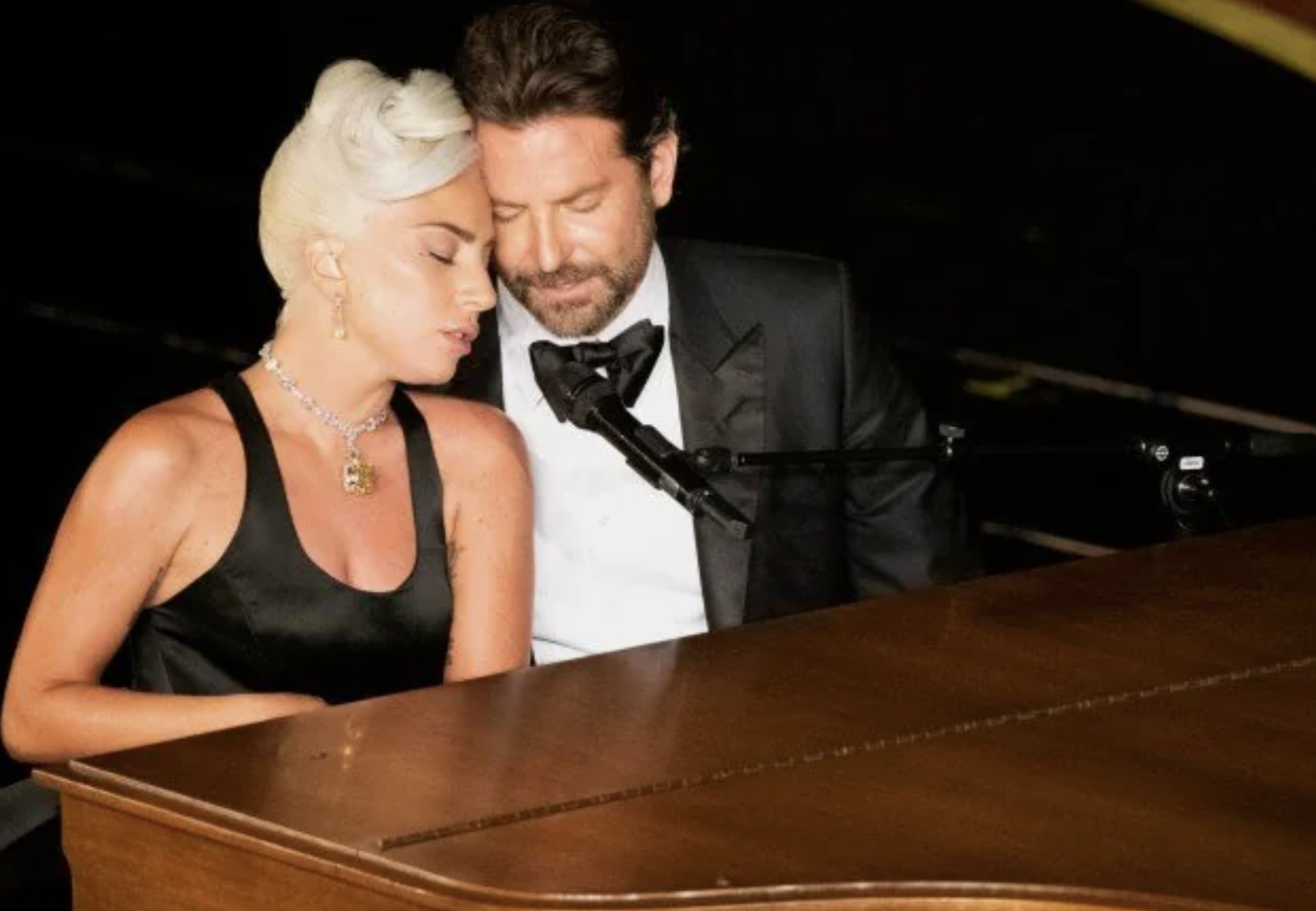 A blonde woman in a black dress and a man in a tuxedo sit closely together at a piano, both appearing to sing with emotion under stage lighting.