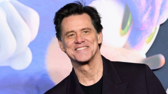 A man with dark hair, wearing a black shirt and jacket, smiles at the camera. The background features a large, colorful illustration of a blue animated character with a white glove.