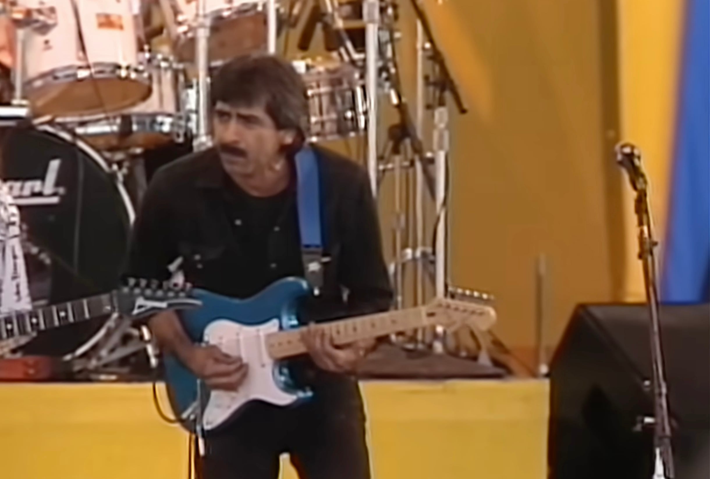 A man with dark hair and a mustache plays an electric guitar on stage. He wears a black shirt and stands in front of a drum set, with yellow and blue stage decorations visible in the background.