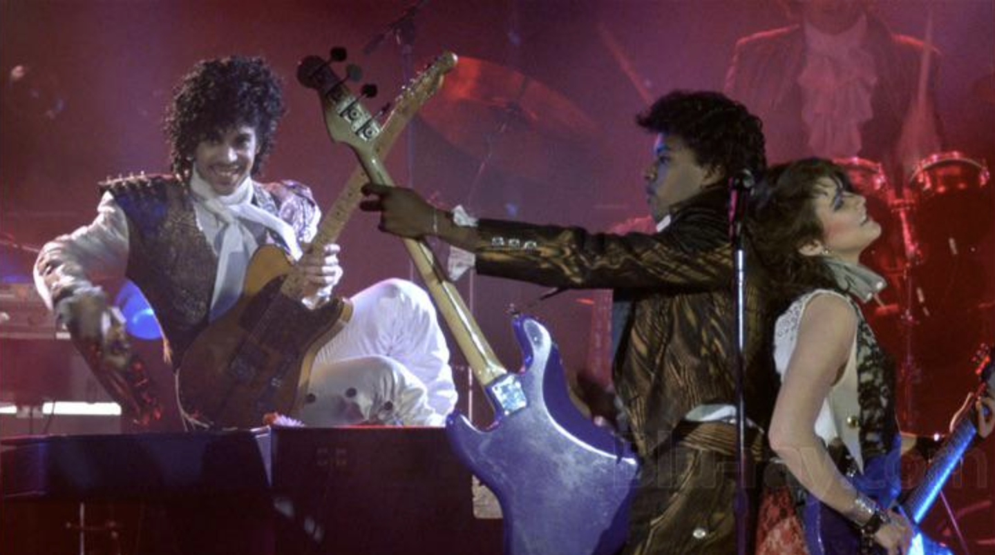 Prince and his band perform energetically on stage with vibrant lighting; Prince smiles while playing keyboard, a guitarist lifts his instrument, and a female band member stands nearby with a microphone.