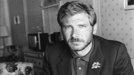 A man with short hair and a beard, wearing a suit jacket and collared shirt with a flower on the lapel, gazes seriously at the camera in a vintage-style, softly lit room.