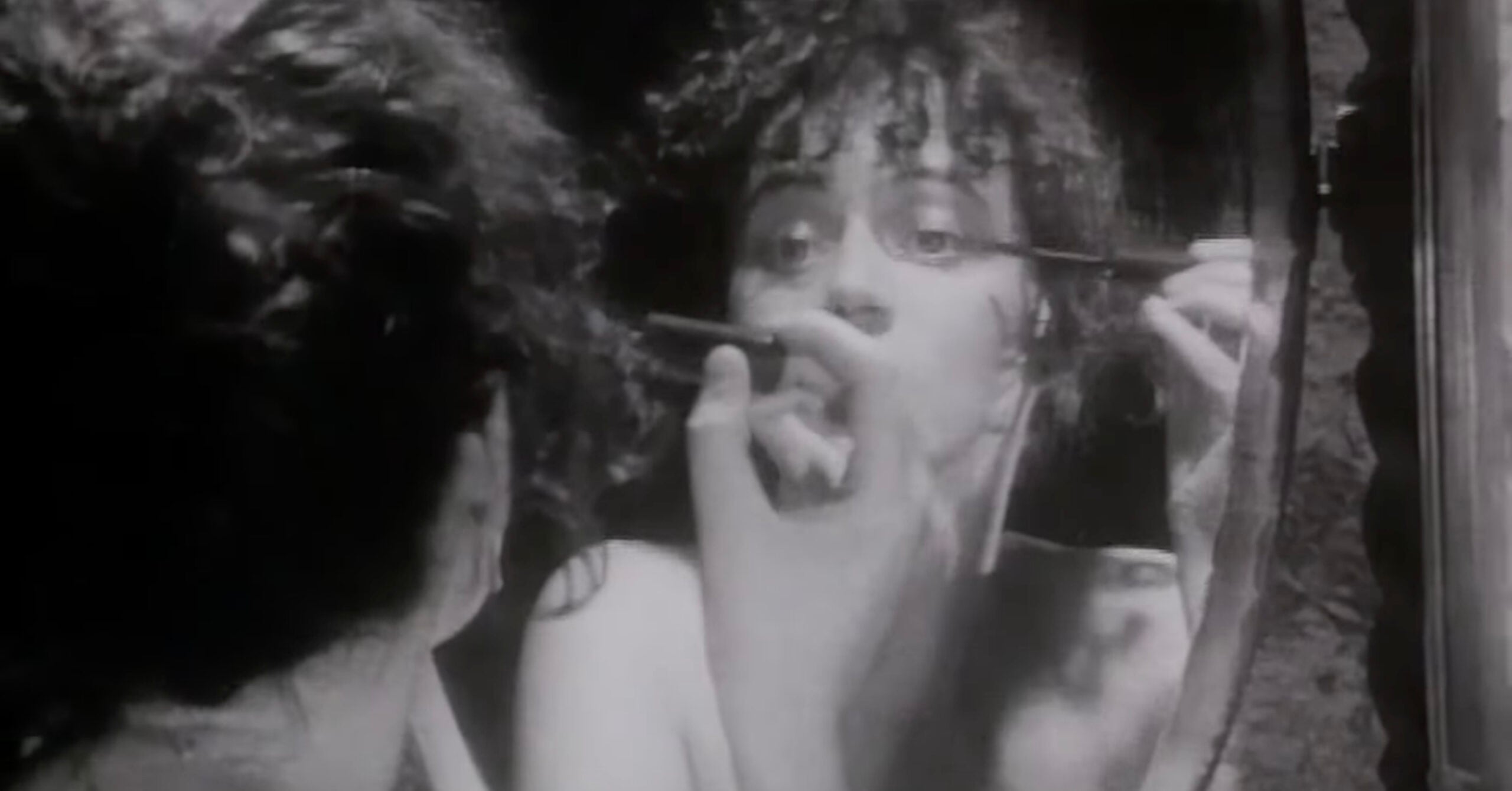 A person with curly hair applies makeup while looking into a mirror, holding an eyeliner pencil near their eye. The image is in black and white and shows the person's reflection clearly.