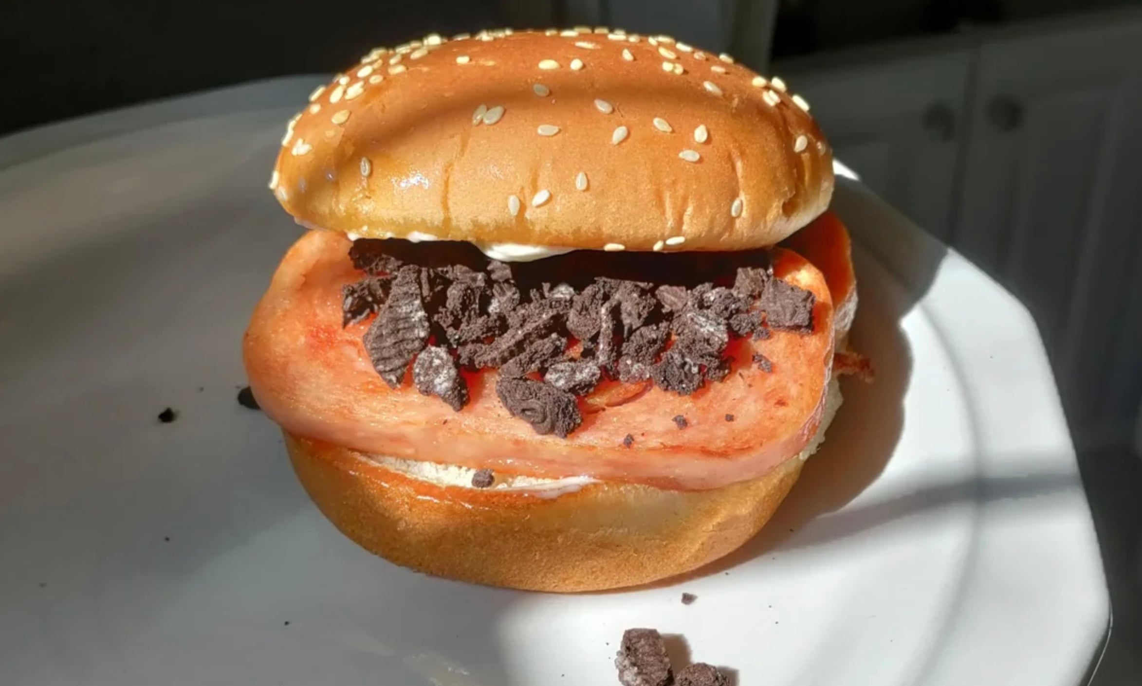 A sesame seed hamburger bun sandwich with a slice of luncheon meat and crushed chocolate sandwich cookies on a white plate, lit by sunlight.