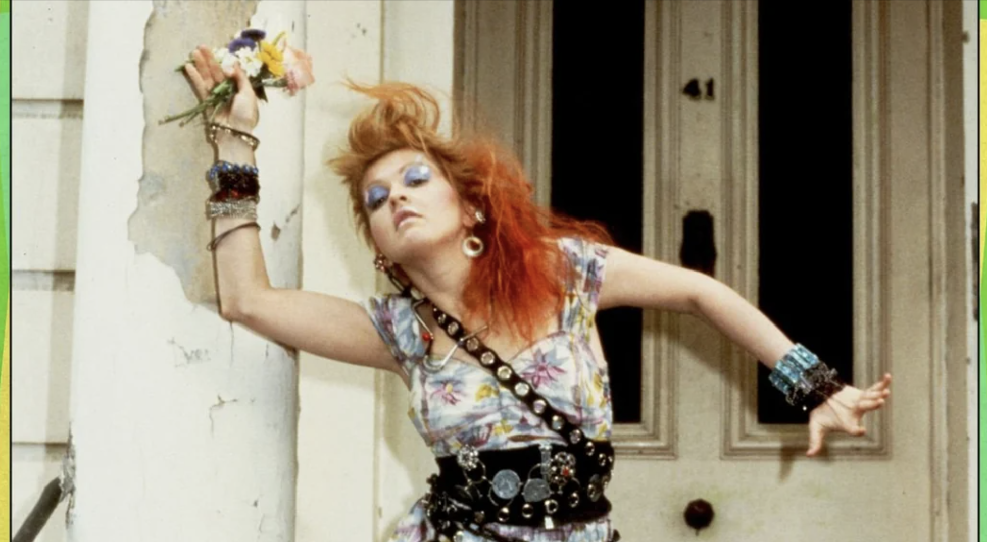 A woman with bright red hair and colorful makeup strikes a dramatic pose against a wall, wearing a floral dress, layered belts, and many bracelets, holding flowers. The background shows a door and part of a white building.