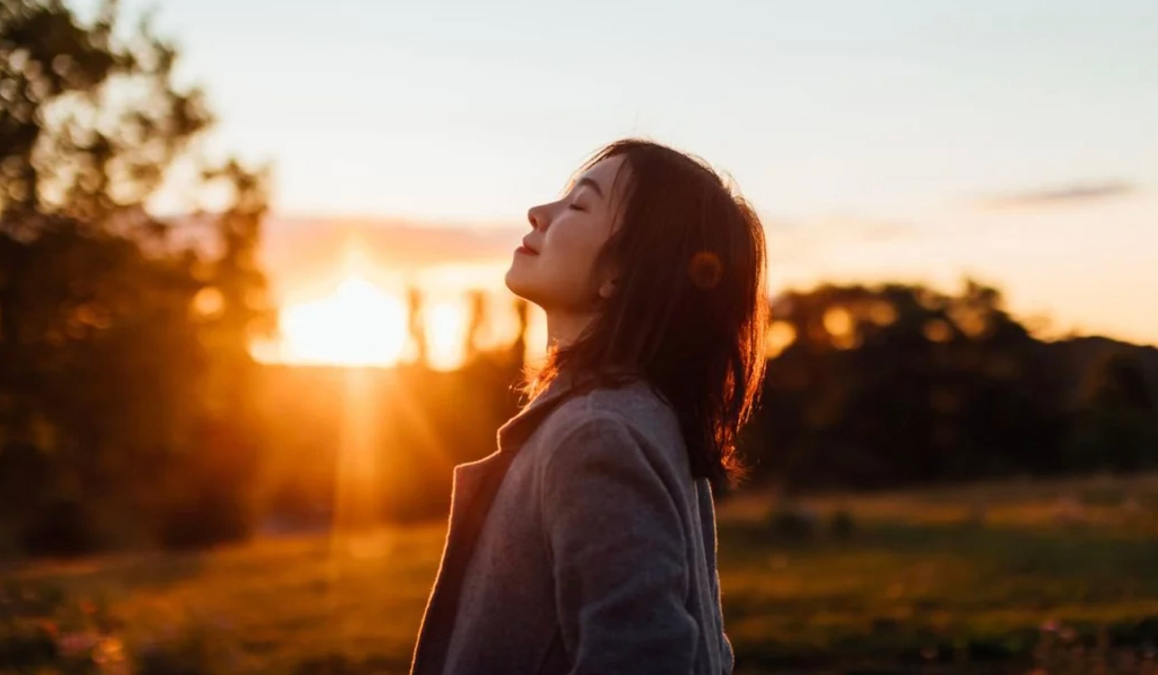 A woman with shoulder-length hair stands outdoors with her eyes closed, facing the sunset. She appears relaxed and is wearing a gray coat, with warm sunlight illuminating her face and the background.