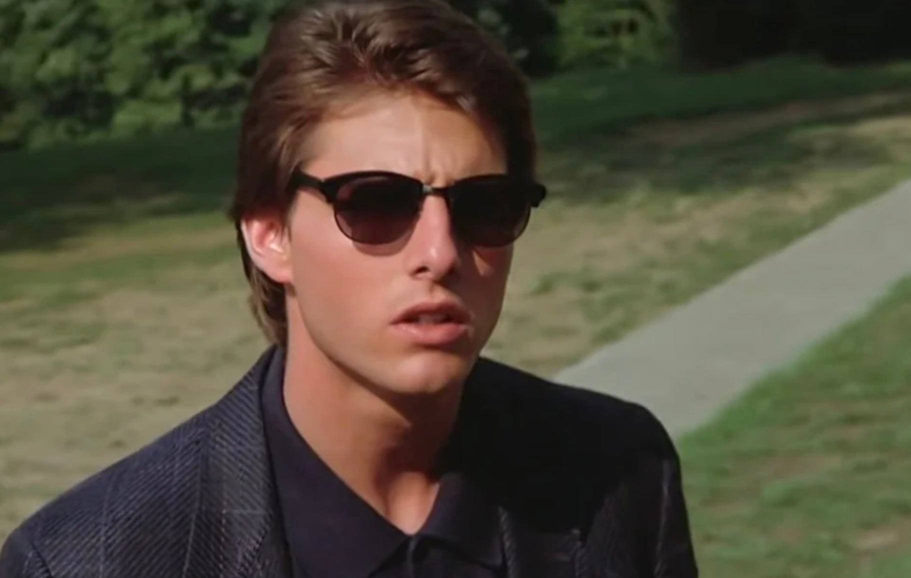A young man with brown hair wearing dark sunglasses and a dark blazer stands outside on a sunny day, with grass and a pathway visible in the background.