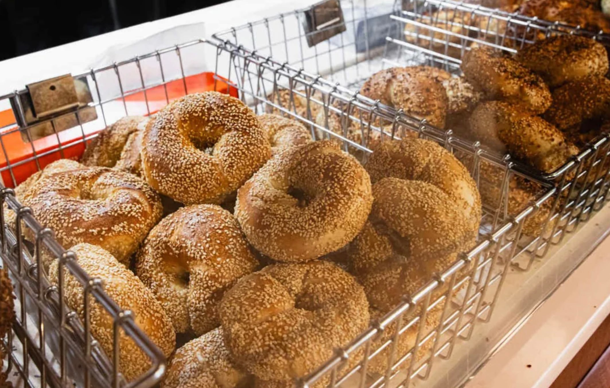 Wire baskets filled with fresh sesame seed bagels are on display at a bakery counter. The bagels are golden brown and stacked closely together.