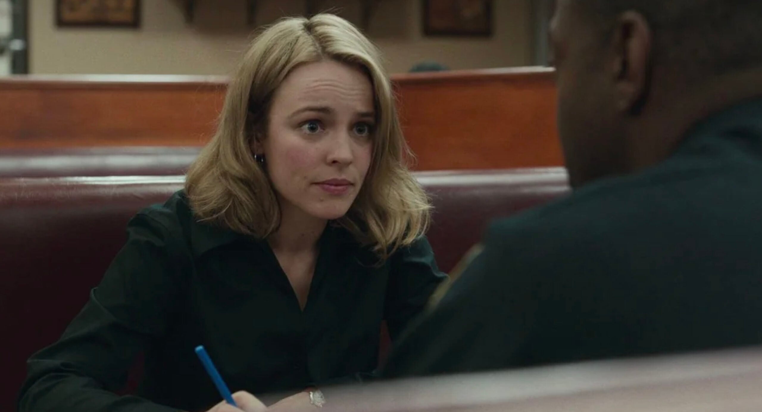 A woman with shoulder-length blonde hair sits in a booth, holding a pen and looking attentively at a person seated across from her. The background shows brown wooden booth dividers.