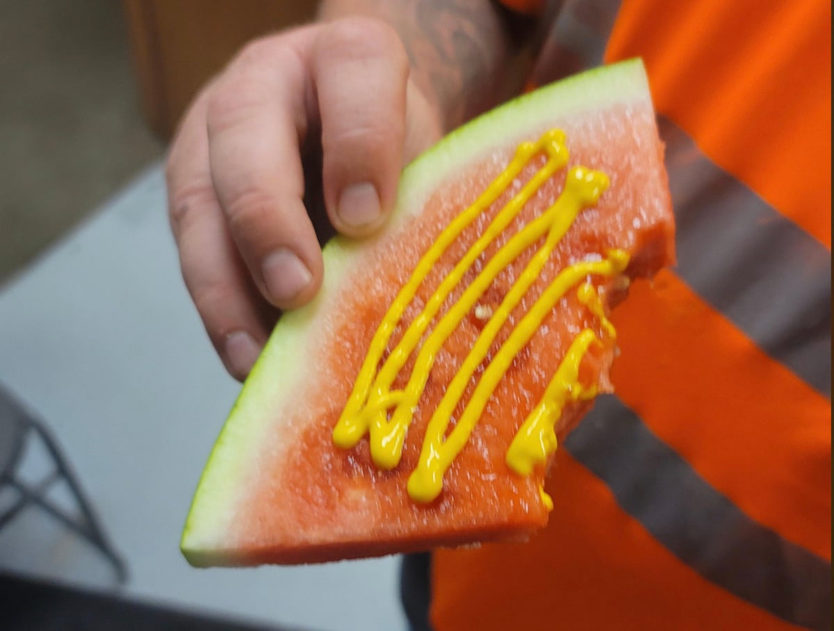 A person wearing an orange shirt holds a slice of watermelon with several lines of yellow mustard squeezed on top.