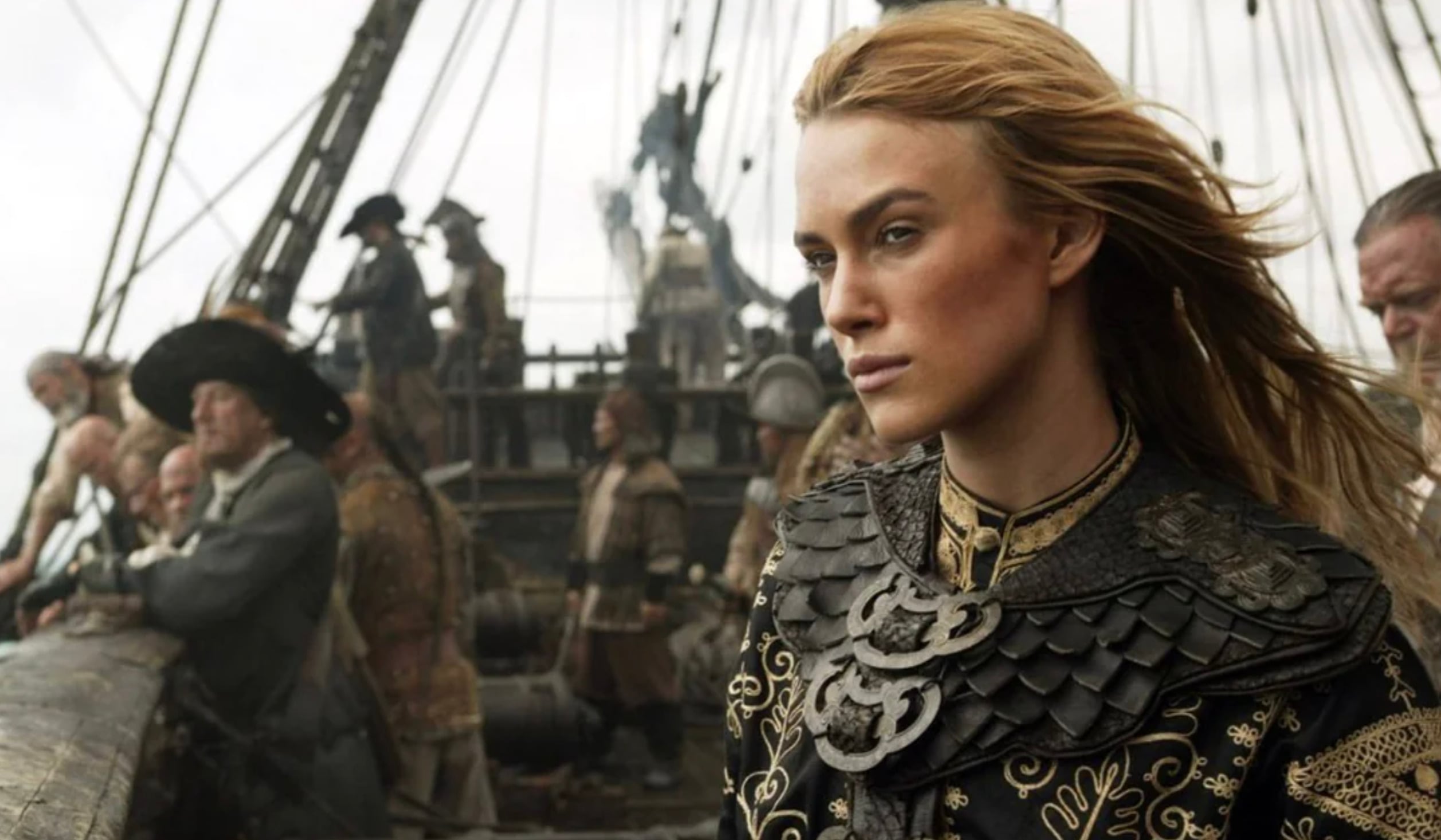 A woman in ornate armor stands on the deck of a ship, looking determined. Behind her, crew members work among ropes and sails. The scene is set on the open sea, with a cloudy sky above.