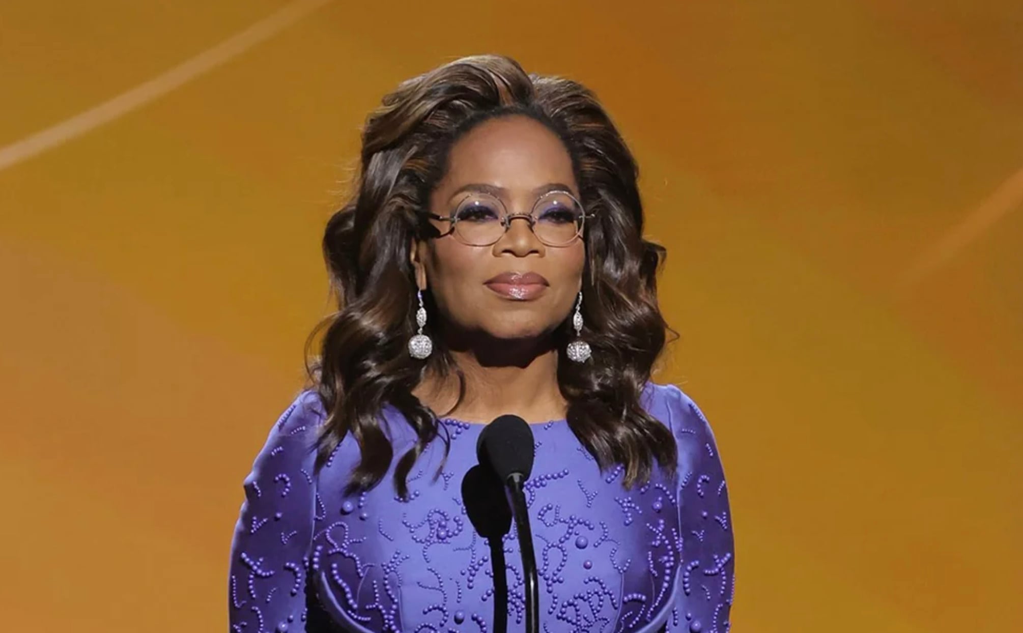 A woman with wavy hair, wearing glasses, drop earrings, and a textured purple dress stands at a microphone against a gold background.