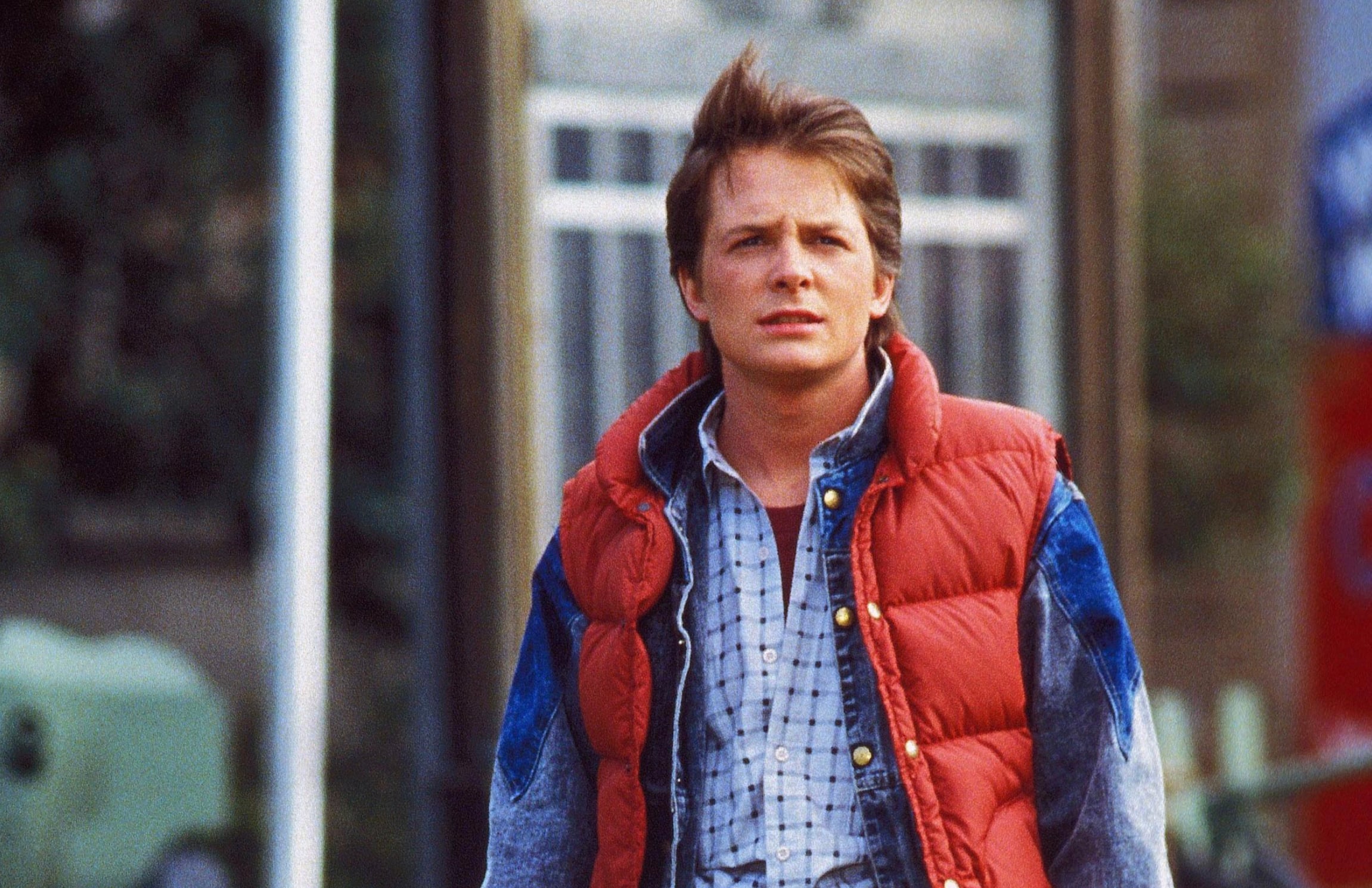 A young man with short brown hair, wearing a red puffy vest over a denim jacket and plaid shirt, stands outdoors in a town setting, looking ahead with a serious expression.
