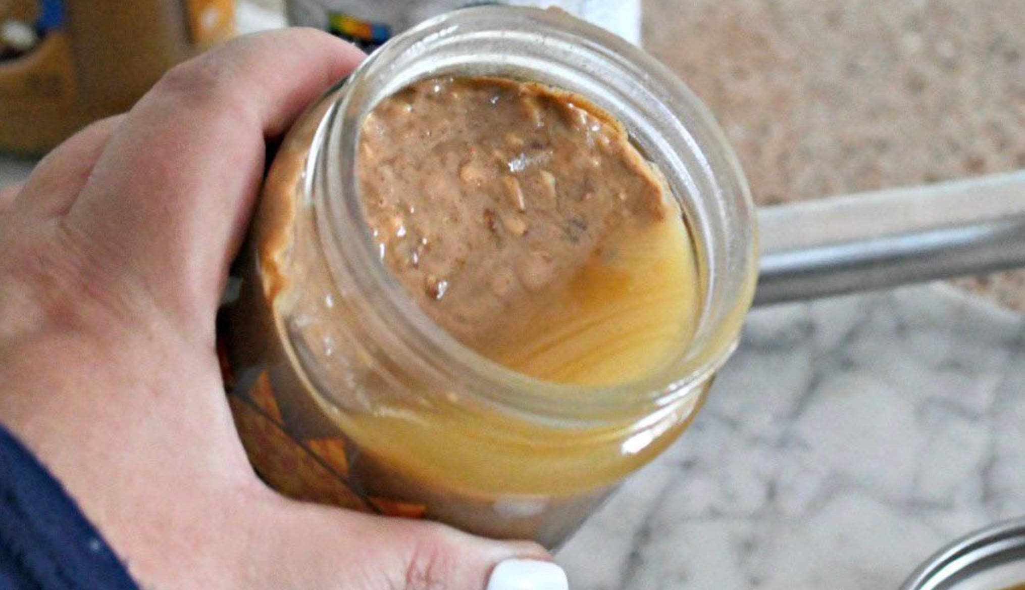 A hand holding an open jar of natural peanut butter, showing separated oil on top of the peanut butter inside the jar.