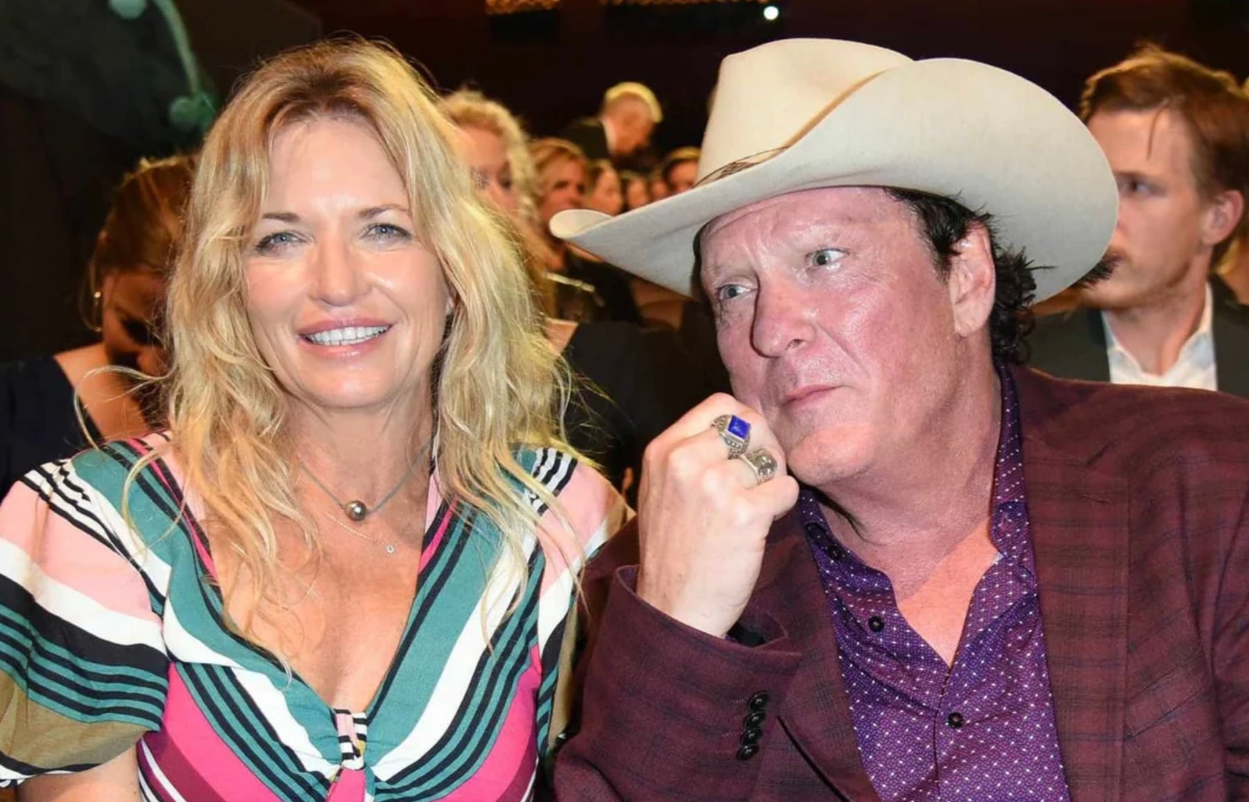A smiling woman in a colorful striped dress sits next to a man wearing a light cowboy hat and a maroon checkered jacket. They are seated among a crowd at what appears to be an indoor event.