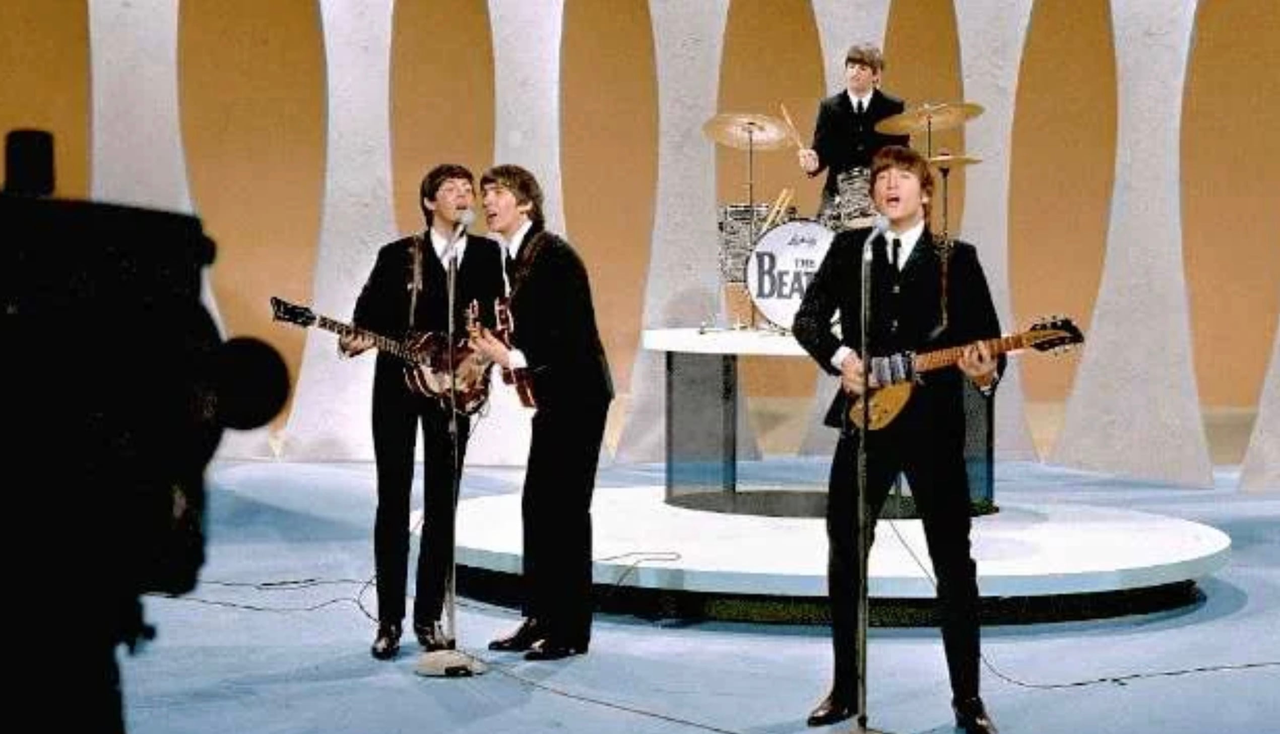 Four musicians in black suits perform on a bright stage, three playing guitars and singing at microphones while one plays drums in the back. The drum kit displays “The Beatles.” A camera is visible in the foreground.