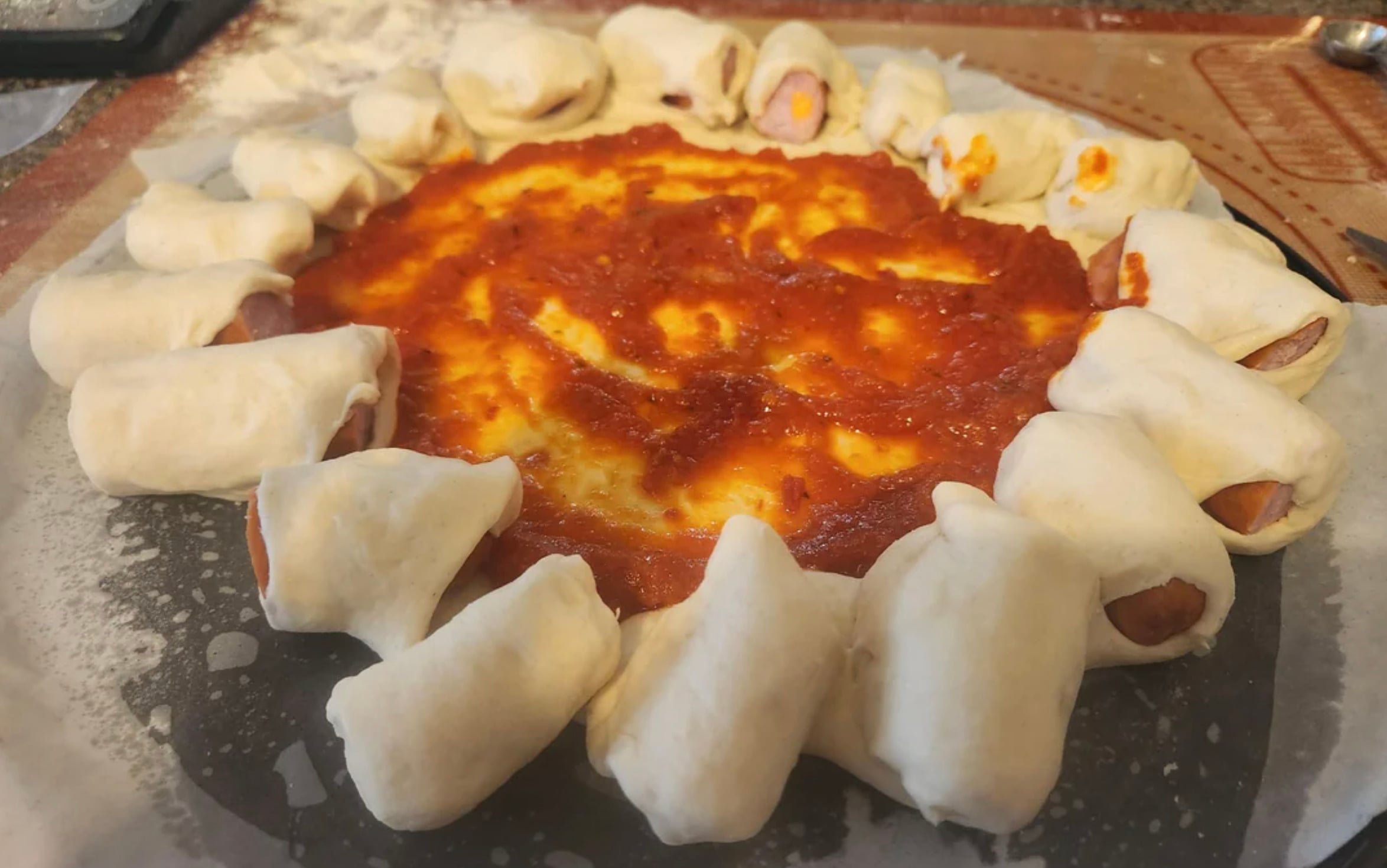 A pizza with a ring of uncooked dough-wrapped mini sausages forming a crust around tomato sauce and melted cheese in the center, on a floured surface before baking.