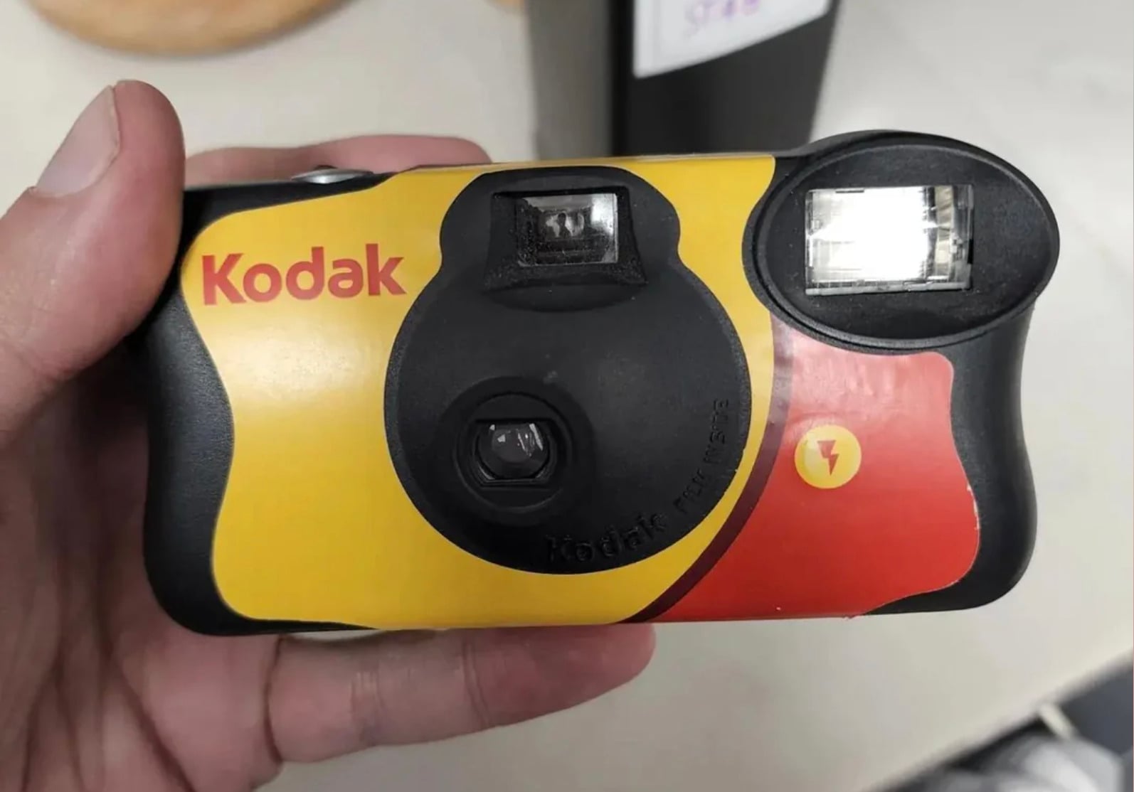 A hand holding a Kodak disposable film camera with a yellow, red, and black design. The camera features a lens, viewfinder, and built-in flash.