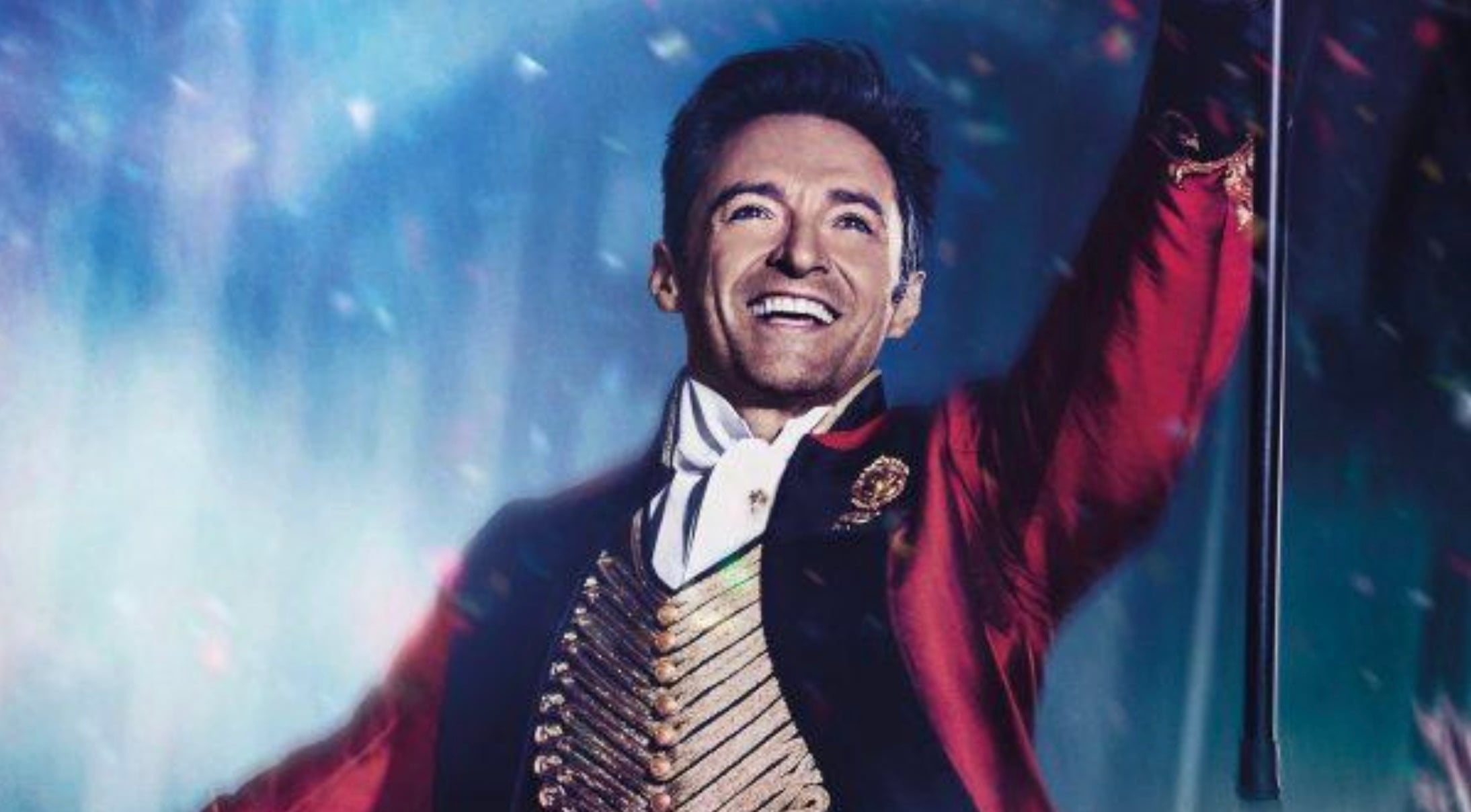 A man wearing a red tailcoat, gold-trimmed vest, and white shirt smiles brightly while holding a cane, standing in front of a sparkling blue and purple background.