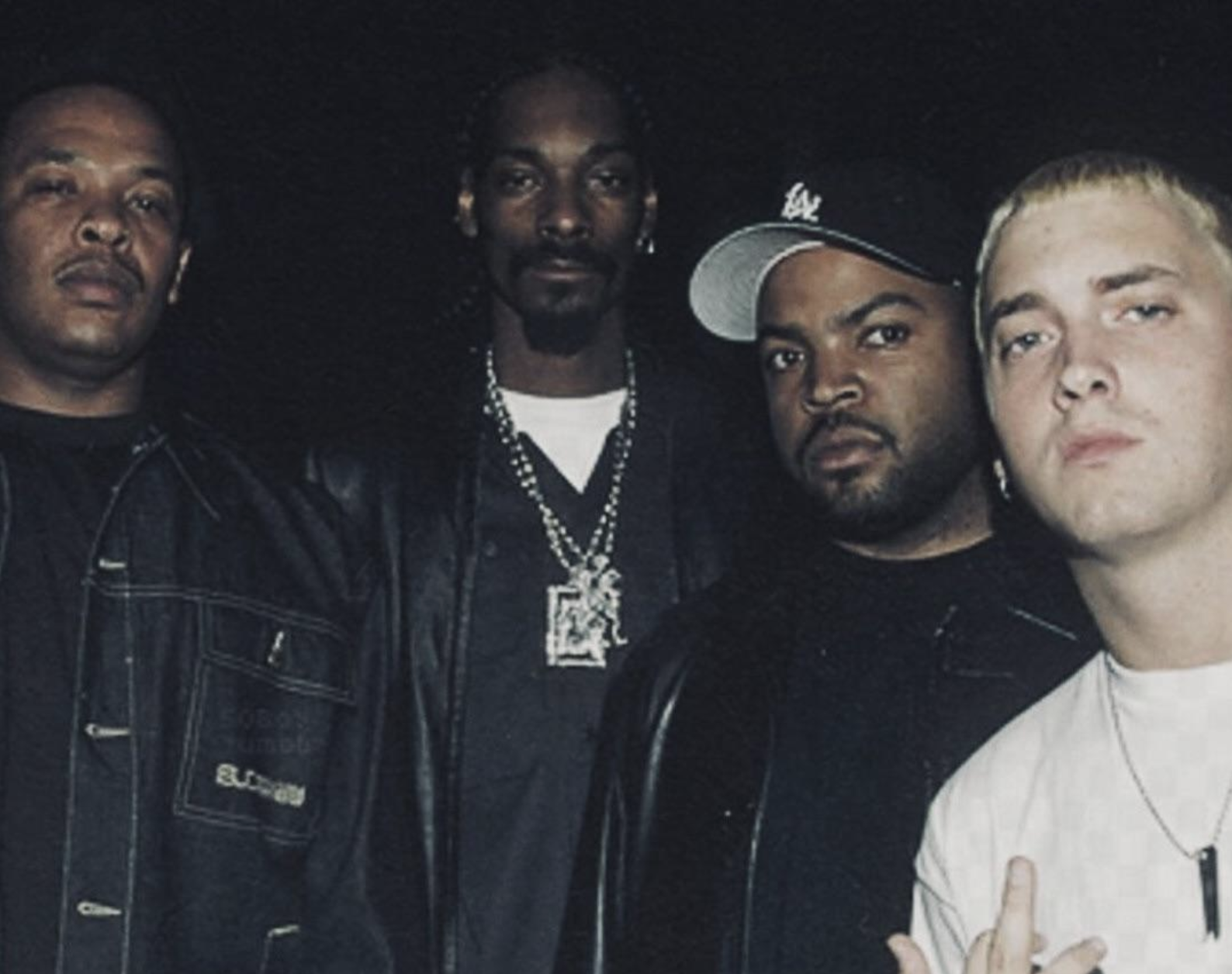 Four men stand together against a dark background, all wearing dark clothing and serious expressions. One man in front is making a hand gesture, while another wears a black cap and chain necklace.