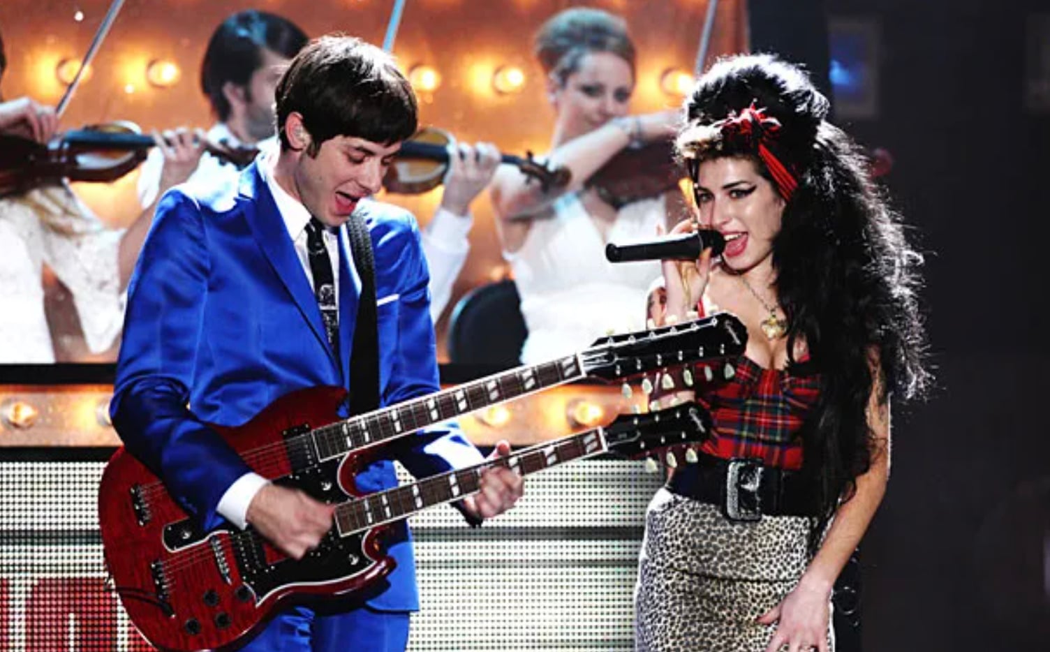 A man in a bright blue suit plays a double-neck guitar next to a woman singing into a microphone, wearing a plaid top and leopard-print skirt, with violinists performing in the background.