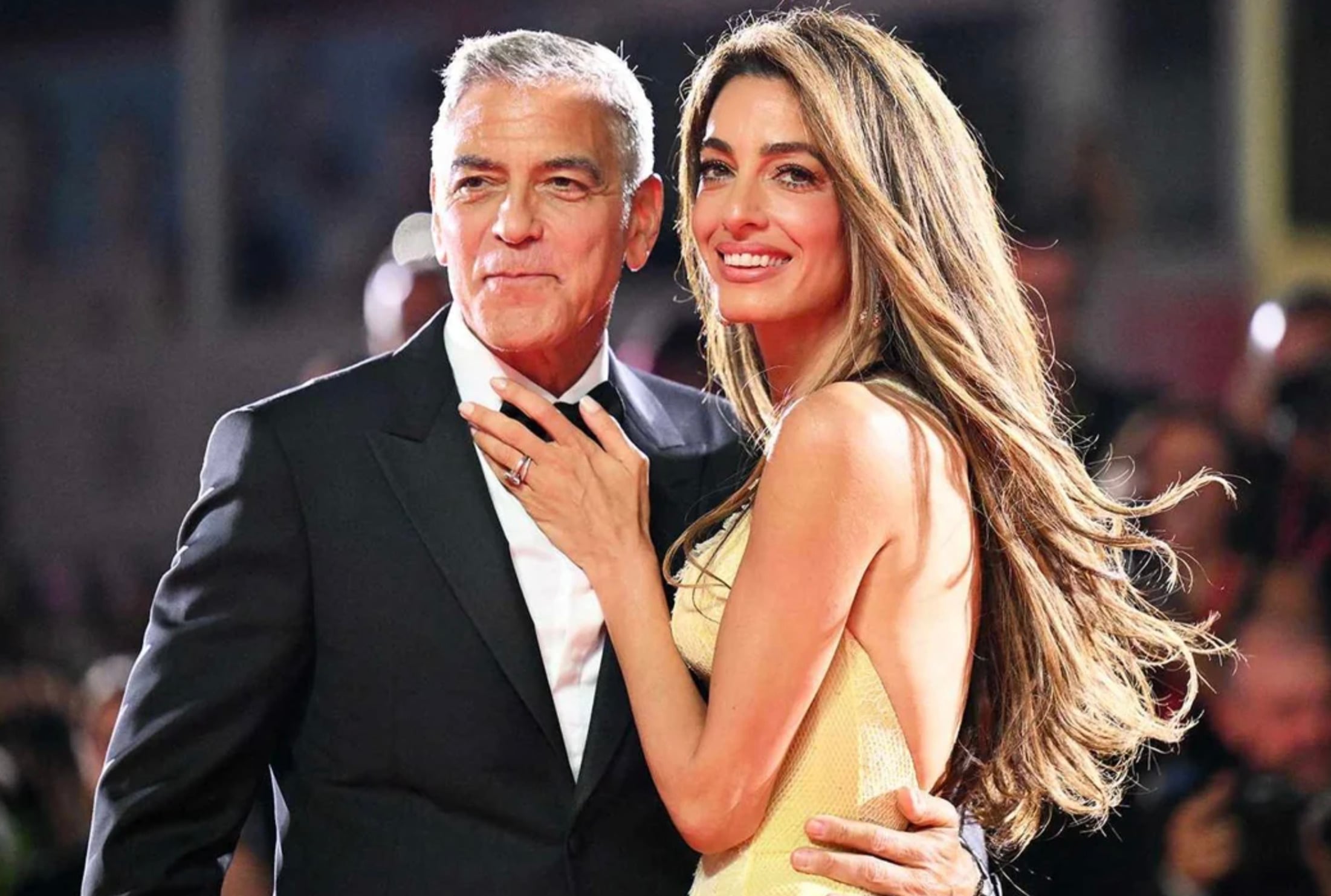 A man in a black tuxedo and a woman in a sleeveless yellow dress smile and pose together on a red carpet at a formal event, surrounded by bright lights and a blurred crowd in the background.