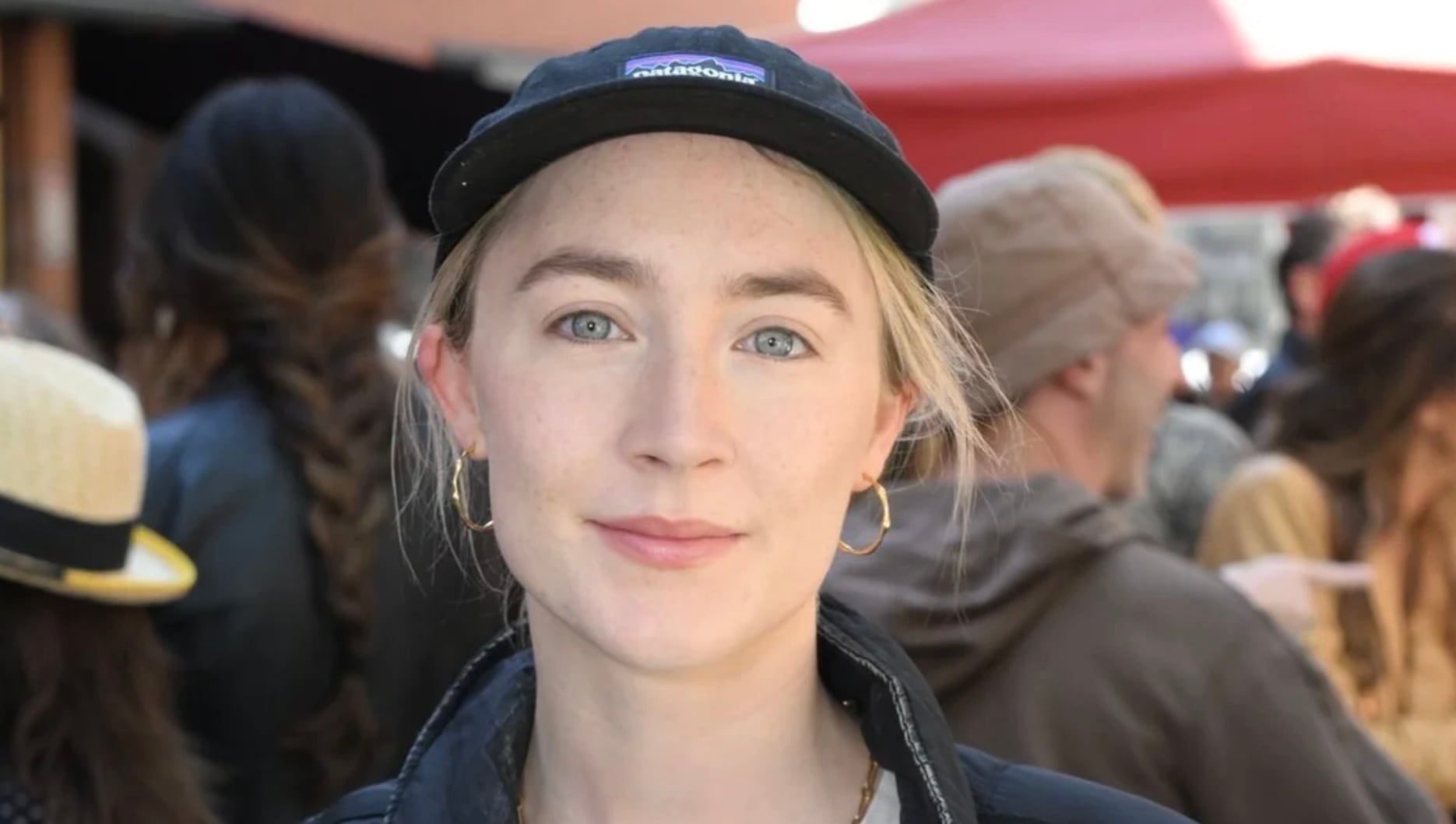 A young woman with light skin, blue eyes, and blond hair wears a black cap and hoop earrings, standing outdoors in a crowd with blurred people and red tents in the background.