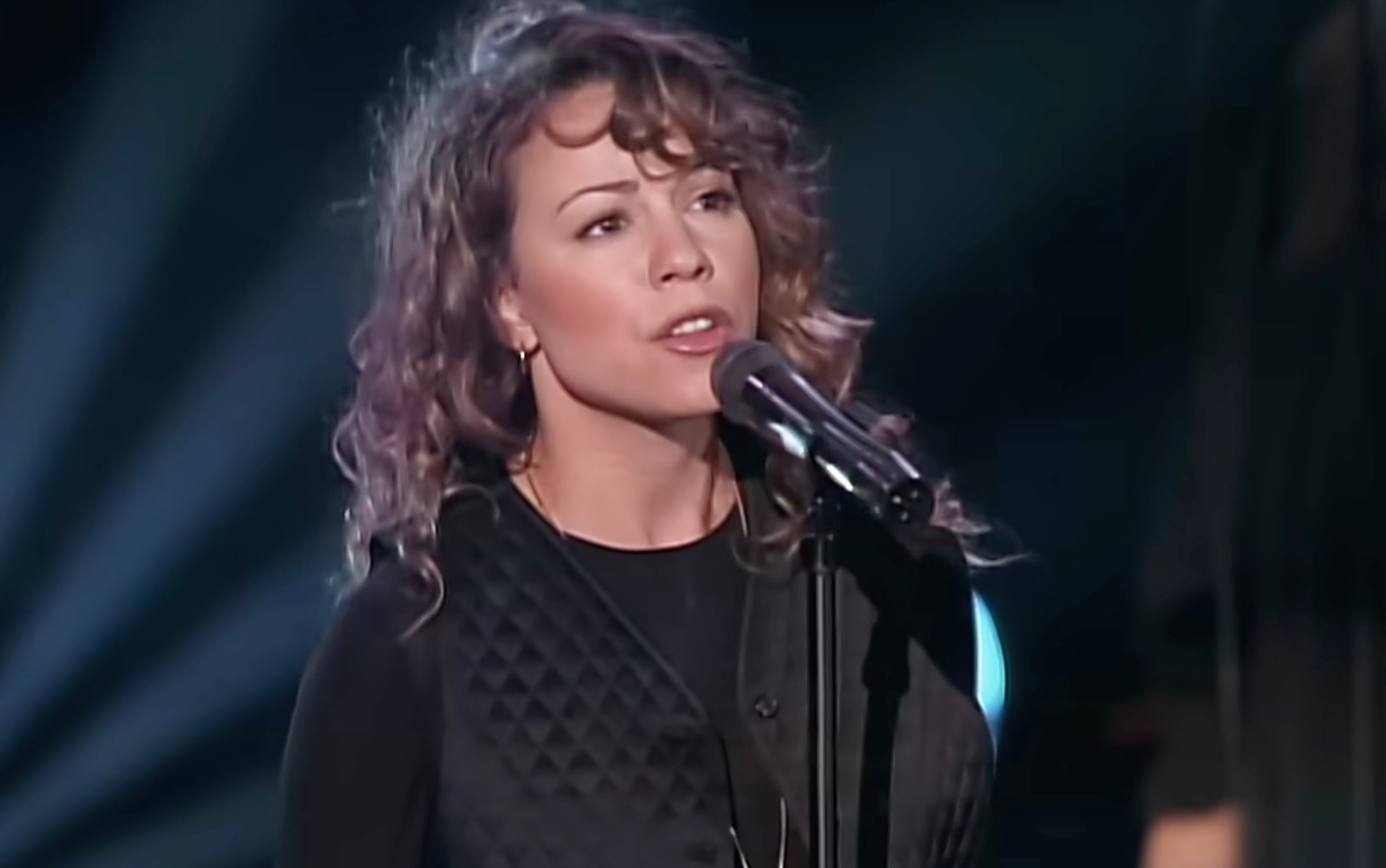 A woman with curly hair sings into a microphone on stage, wearing a black outfit. Stage lights create a dramatic background.