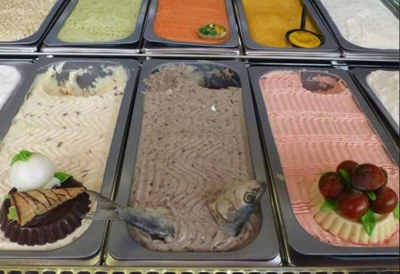 A display of unusual ice cream flavors in metal tubs, some featuring savory ingredients like a fish head, onion, and grapes as decoration, suggesting unique or unconventional tastes.