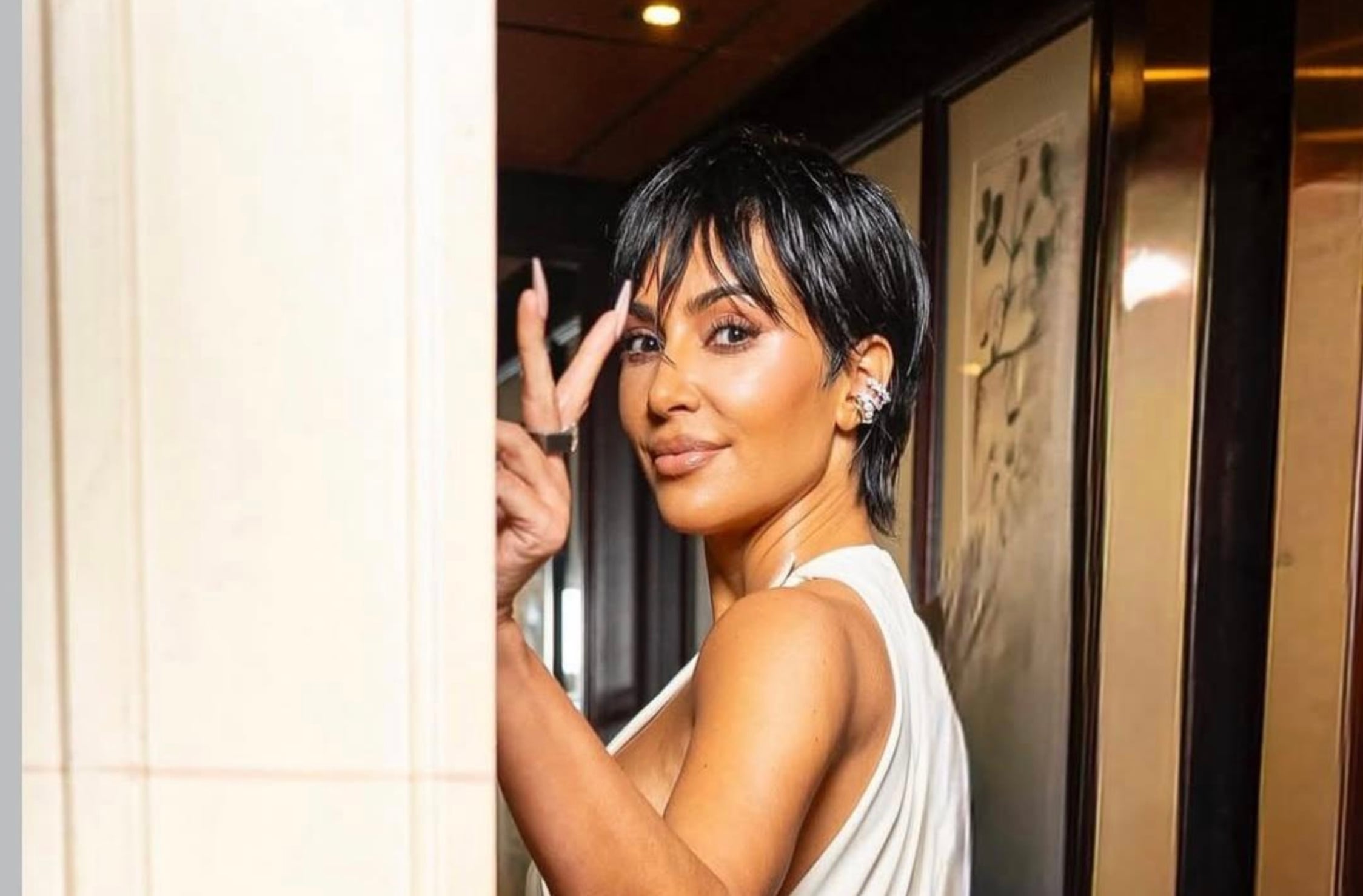 A woman with short dark hair wearing a sleeveless white top stands indoors, smiling and making a peace sign with her hand. She is wearing earrings and posing near a wall with framed art in the background.