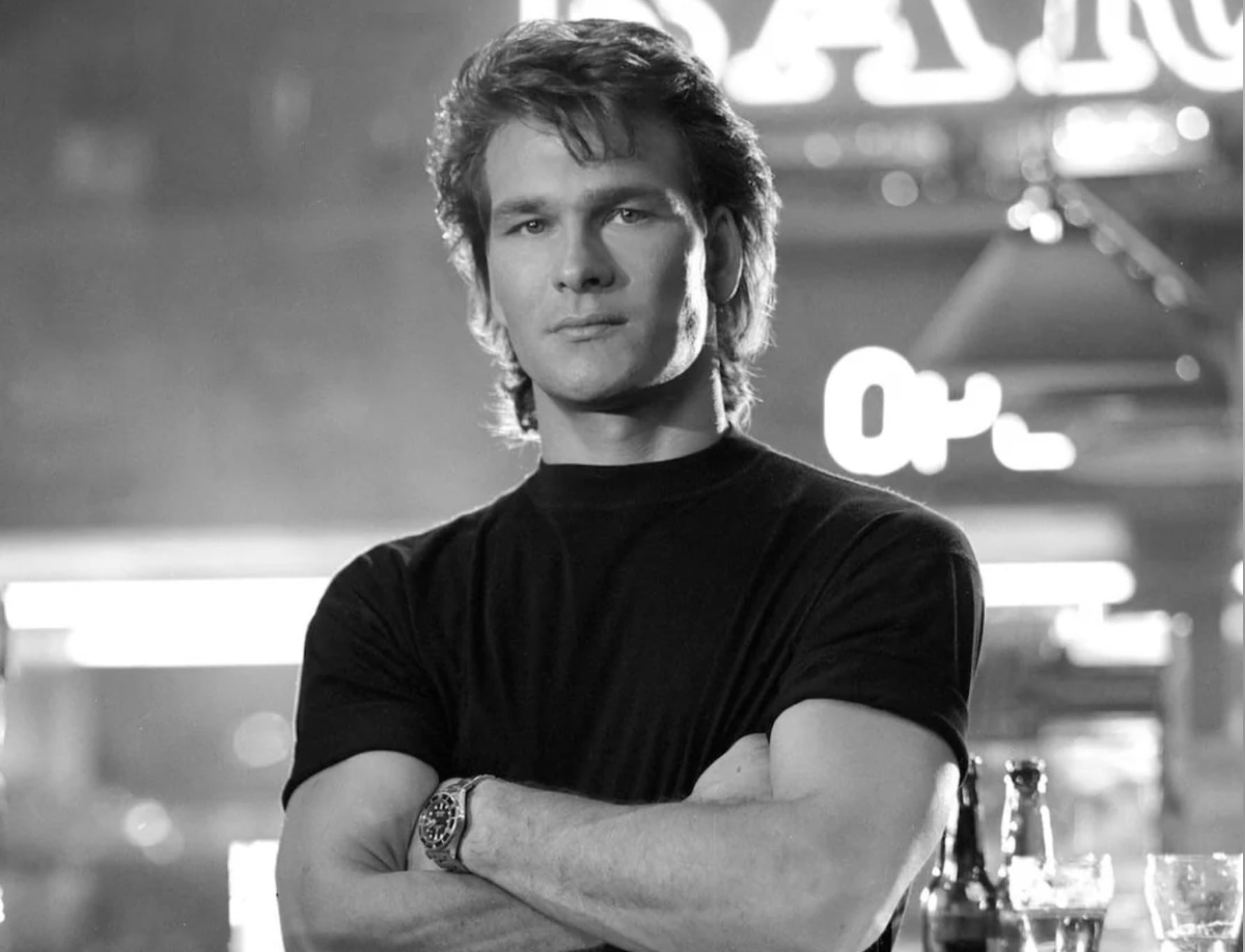 A man with wavy hair wearing a black t-shirt stands with arms crossed in front of a neon-lit bar, looking confidently at the camera. Several bottles and glasses are visible on the bar in the foreground.