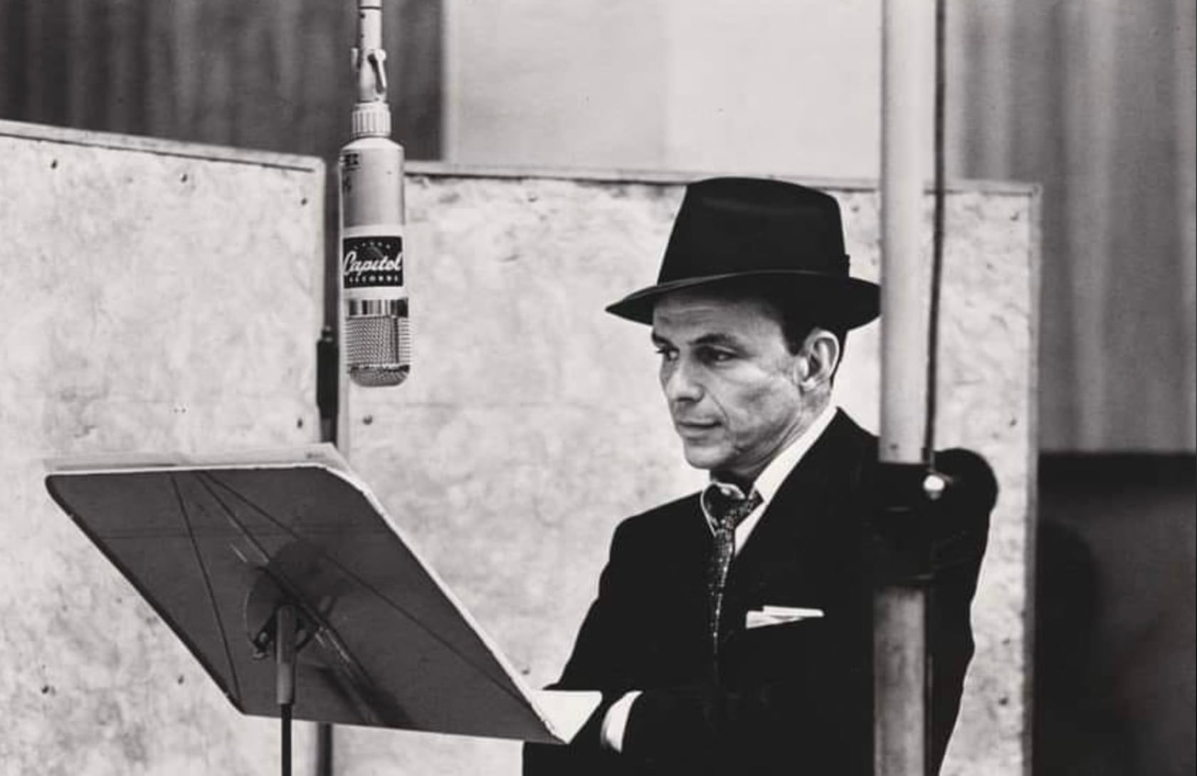 A man in a suit and fedora stands at a music stand in a recording studio, facing a vintage microphone, appearing focused and contemplative.