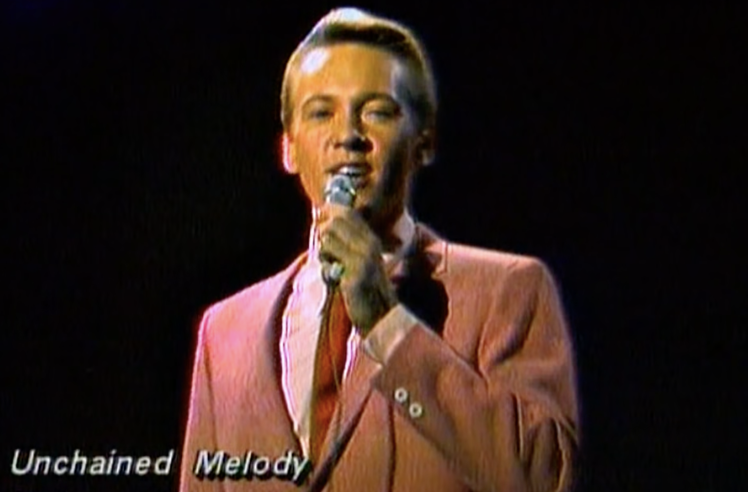 A man in a pink suit and red tie sings into a microphone on stage, with the text "Unchained Melody" displayed in the lower left corner. The background is dark.