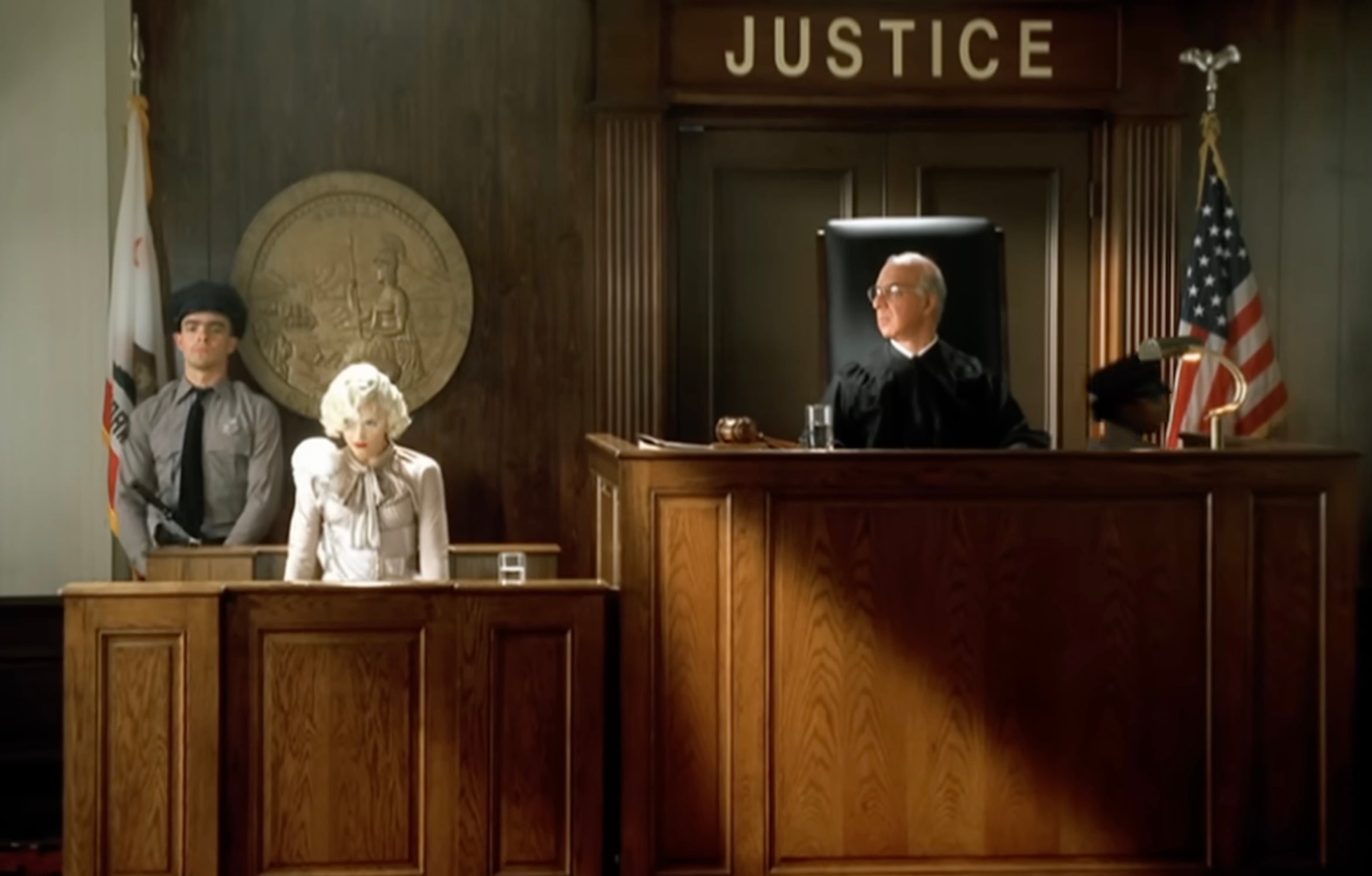 A courtroom scene shows a judge at the bench, a woman in white seated at the witness stand, a man in gray standing nearby, and the word "JUSTICE" above them. An American flag and state seal are visible in the background.
