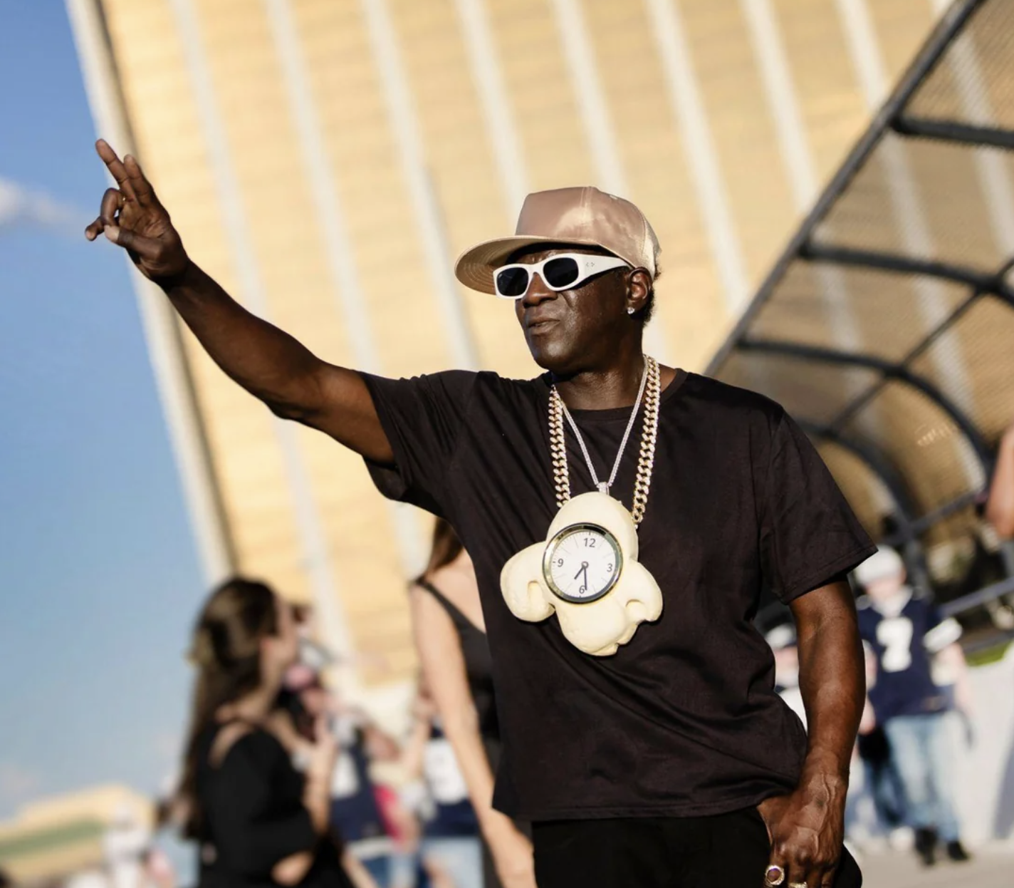 A man wearing a large clock necklace, white sunglasses, and a gold cap poses with a peace sign outdoors; people and tall gold buildings are blurred in the background.