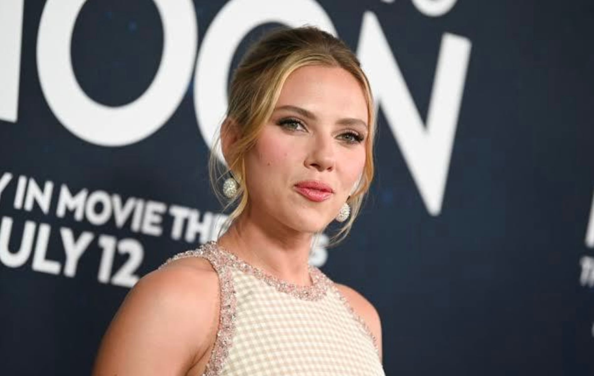 A woman with blonde hair in an elegant updo poses in front of a dark backdrop with white text. She is wearing a sleeveless, light-colored dress with pearl details and drop earrings, and is looking slightly to the right of the camera.