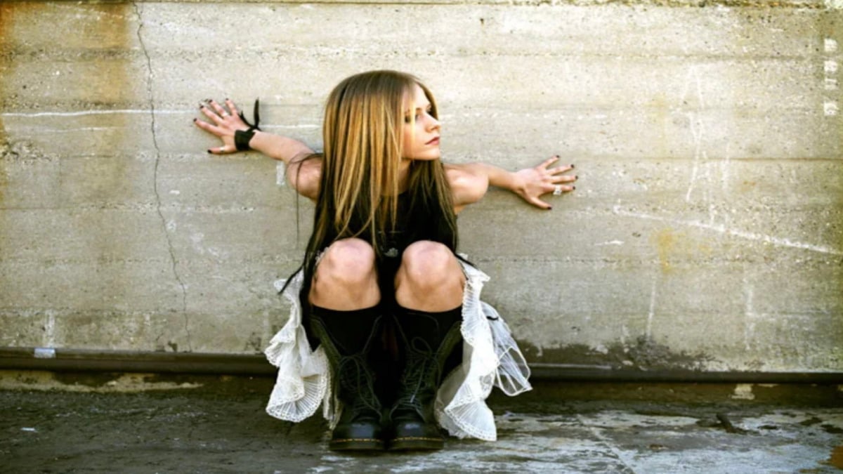 A woman with long blonde hair, dressed in a black top, white skirt, and black boots, sits crouched against a rough concrete wall, spreading her arms to the sides and looking to her left.
